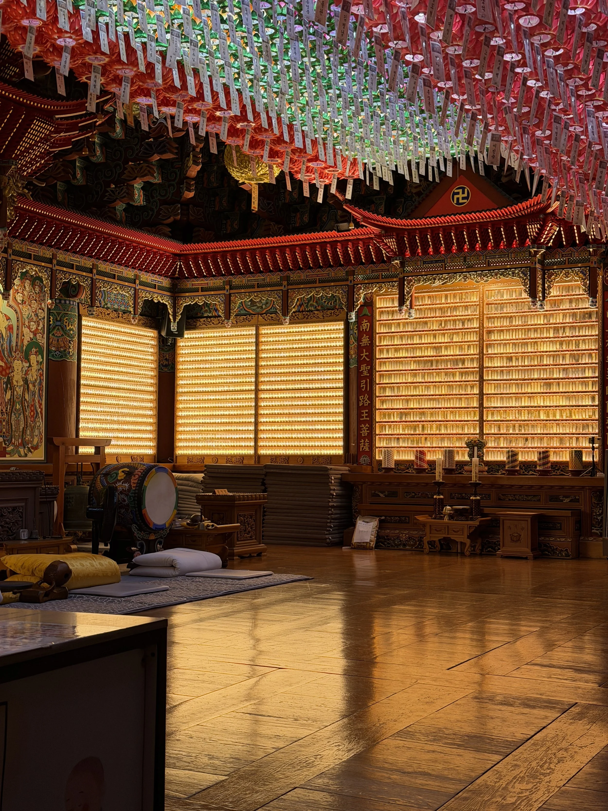 Interior of a Buddhist temple or shrine with ornate decorations, lanterns hanging from the ceiling, painted murals, and musical instruments such as a drum and wooden plaques with Chinese characters.