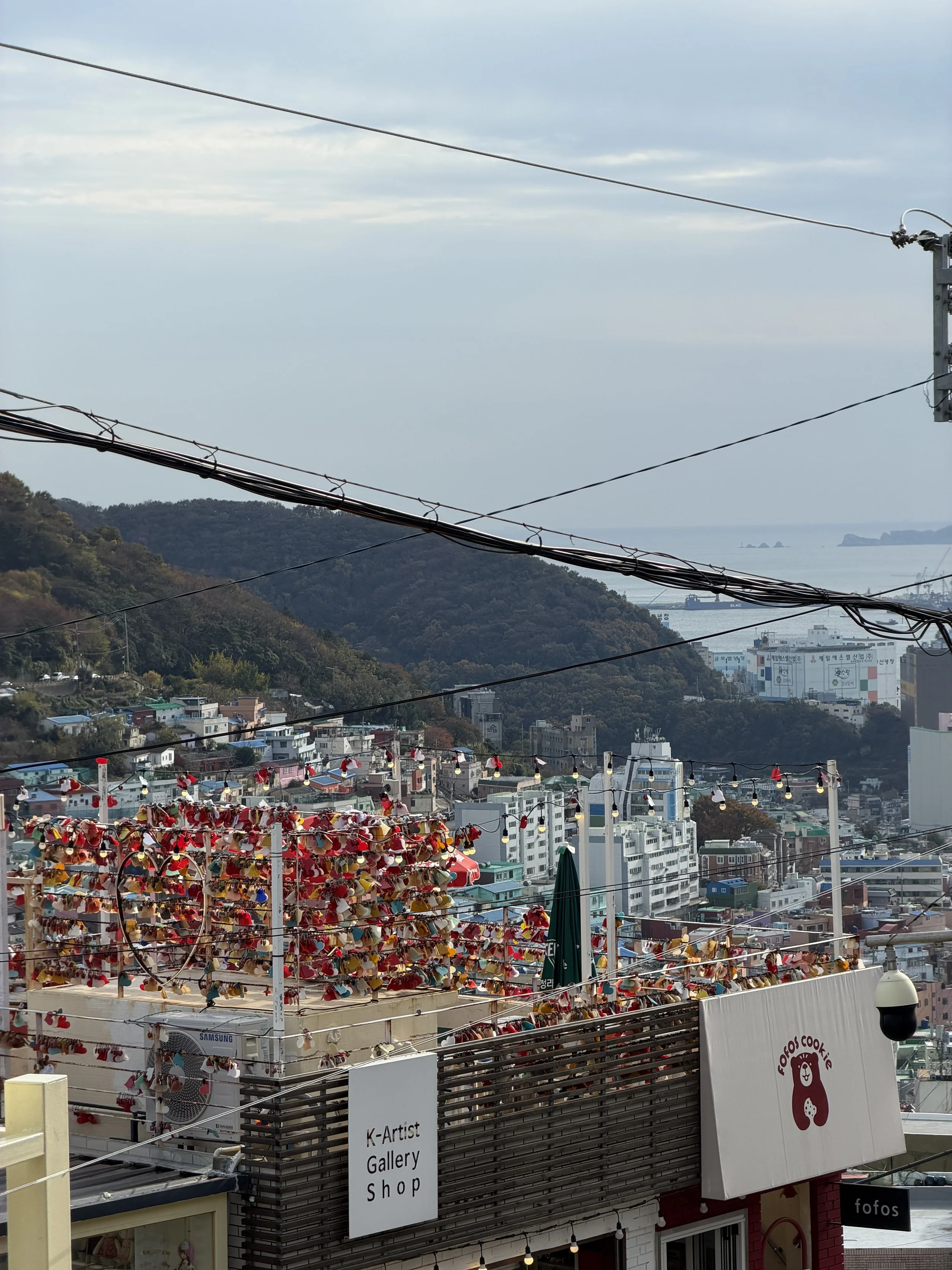 View of a city with hills, buildings, and the sea in the background. In the foreground, covered with colorful hats, is an outdoor display area. Utility wires cross the scene, and there are signs for shops including a 'K-Artist Gallery Shop' and 'fofo