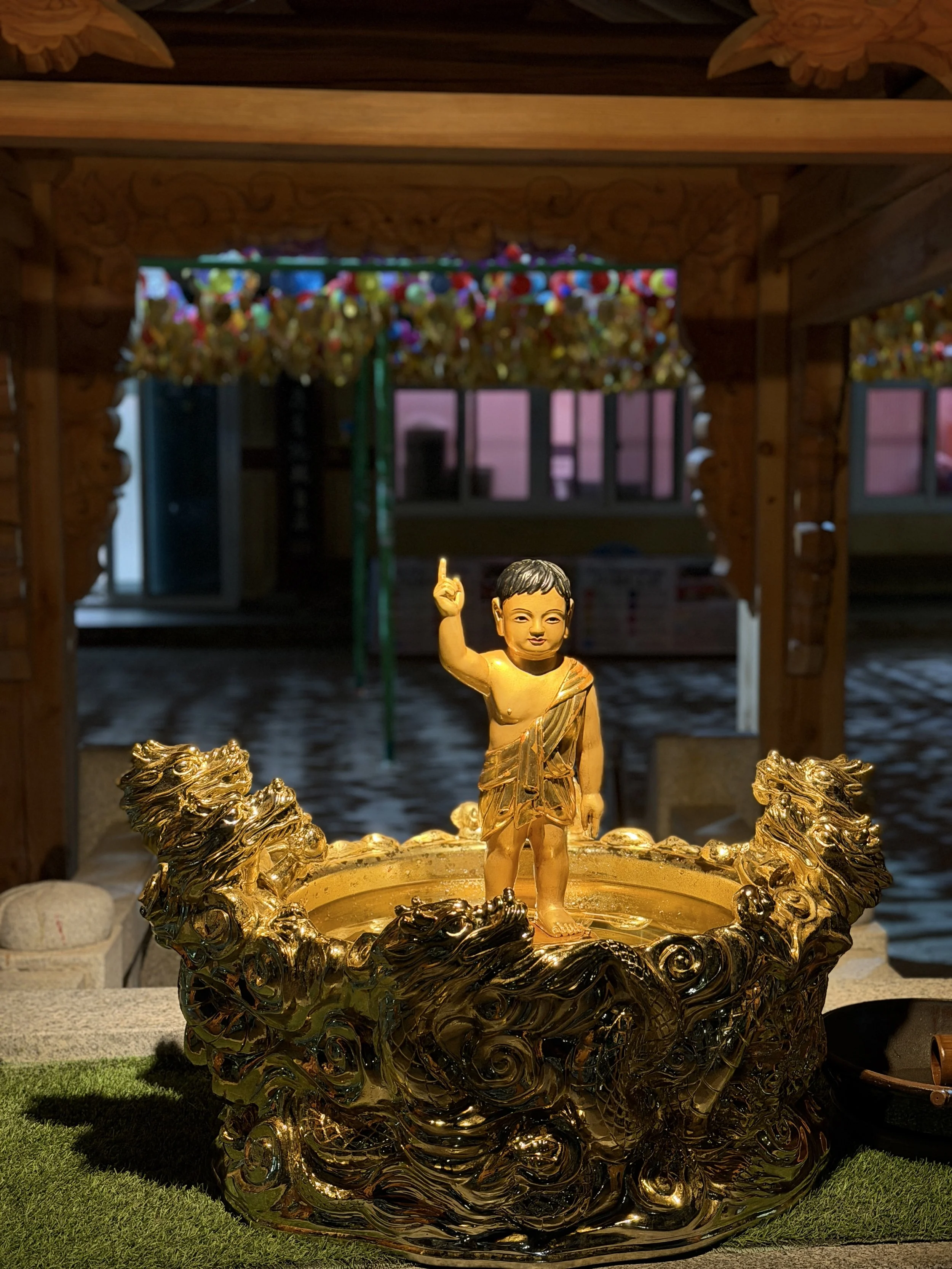 Golden statue of a child in a traditional outfit, standing with one finger raised, inside a decorative golden vessel with dragon motifs, under a wooden structure.
