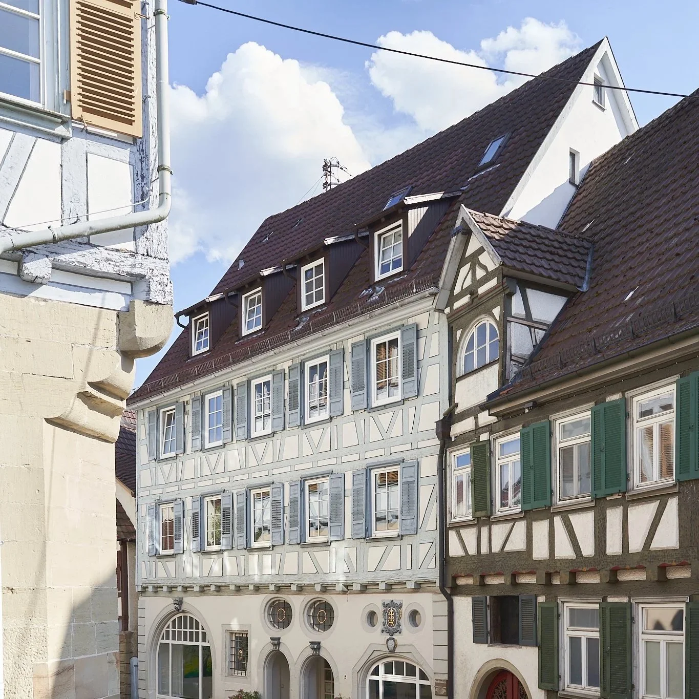 Fassade eines Hauses mit mehreren Fenstern und offenen Holzladenläden, ein Klettergerüst oder Arbeitsbühne mit Seil hängt an der Seite, der Himmel ist blau.