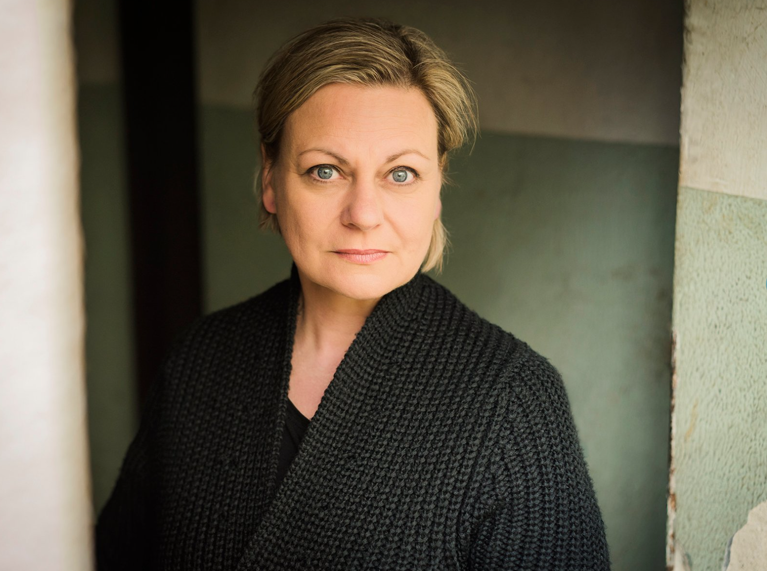 Nahaufnahme einer Frau mit kurzem, hellbraunem Haar, blauen Augen und schwarzer Strickjacke, stehend vor einer grünen Wand mit Putzrissen.