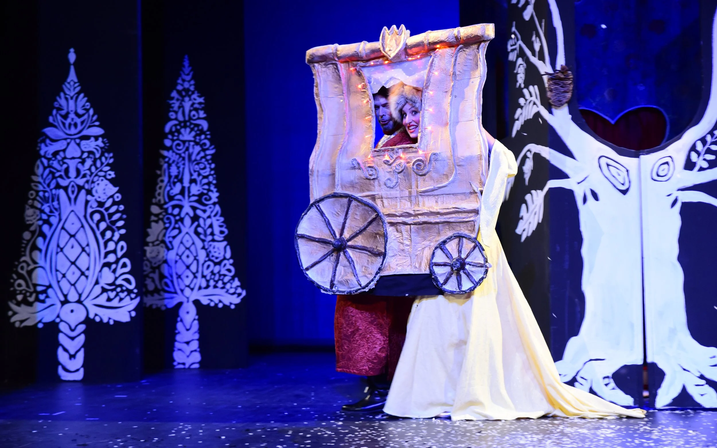 Bühnenaufnahme eines Theaterstücks mit zwei Schauspielern, die in einem Wagen aus buntem Stoff verborgen sind, der als Bustafel dient, vor einem bunten Bühnenhintergrund mit weißen Baum- und Schneemotiven.