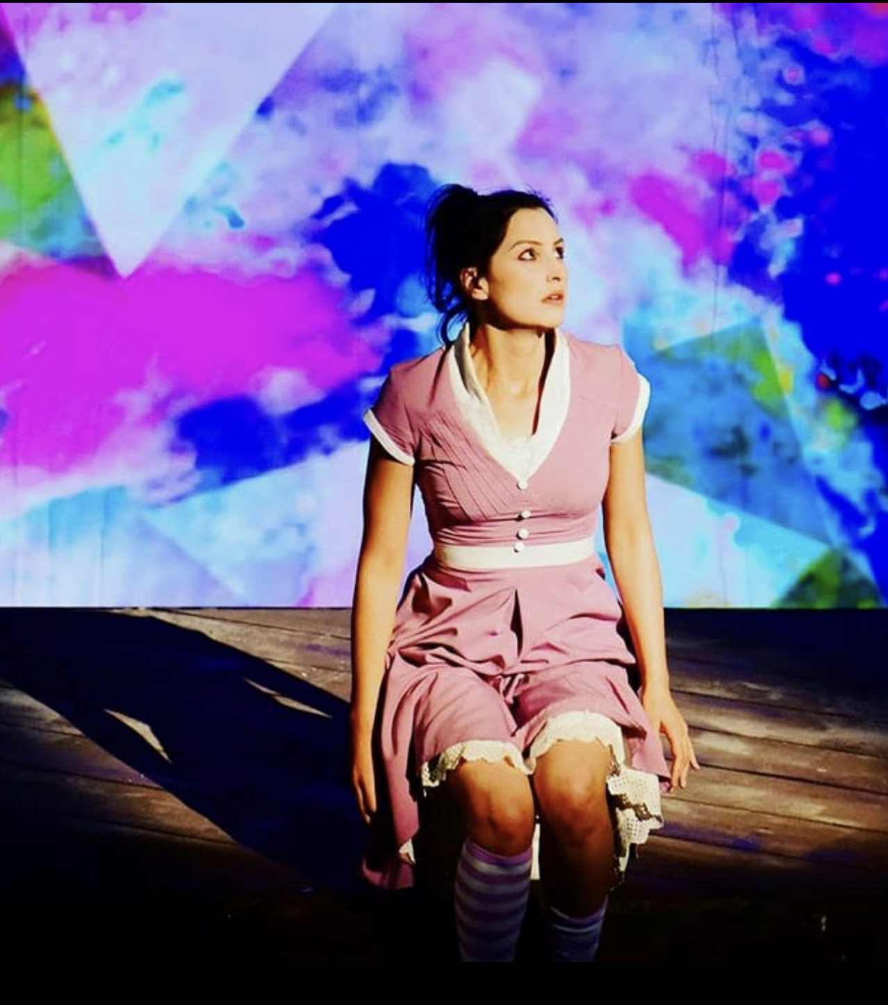 Junge Frau in rosa Kleid mit weißen Details sitzt auf einer Bühne mit bunter, abstrakter Hintergrundprojektion.