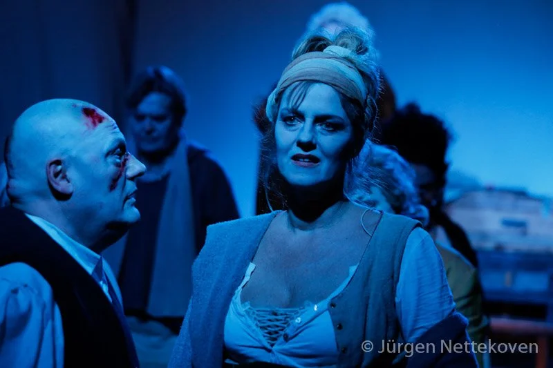 Menschen in einer dramatischen Szene, eine Frau mit Protestoutfit und ein Mann mit blutender Kopfwunde, im Hintergrund weitere Personen, beleuchtet mit bläulichem Licht.