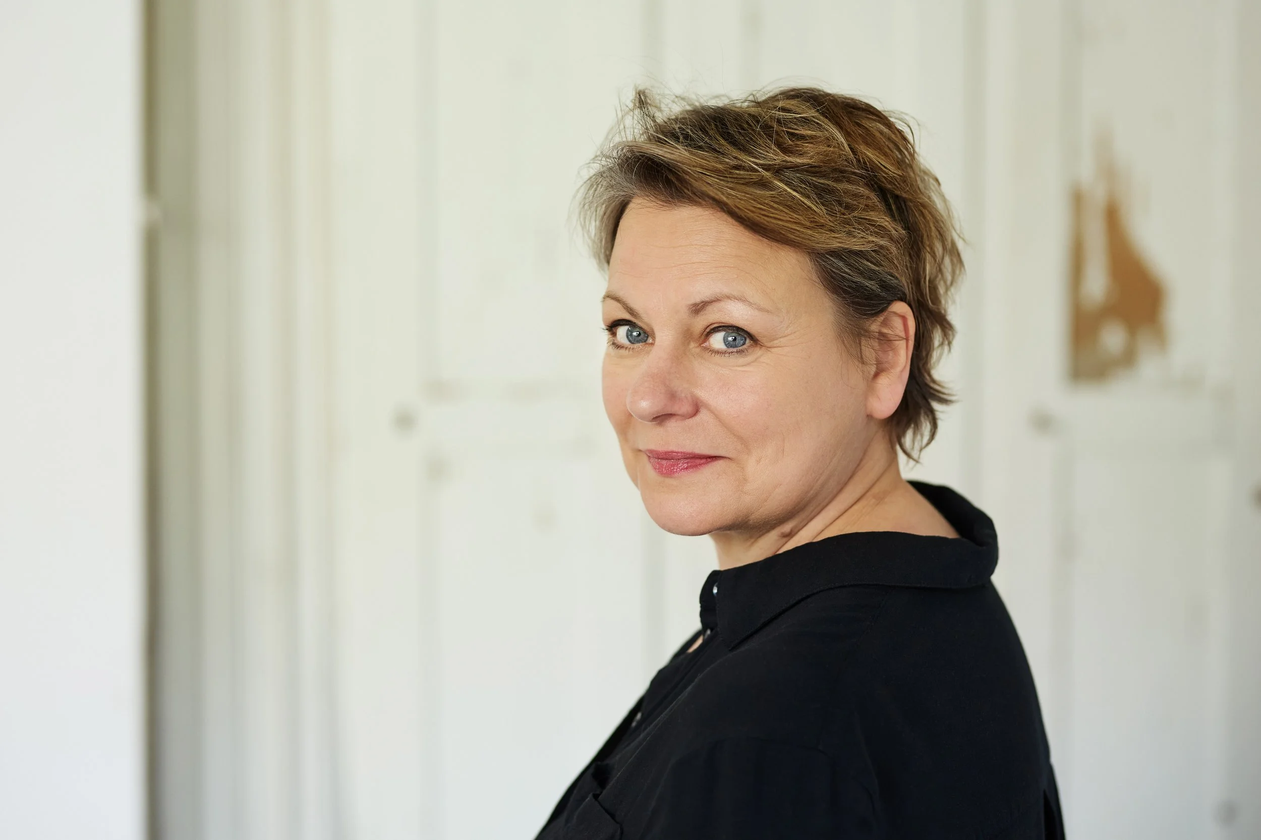 Nahaufnahme einer Frau mit kurzen braunen Haaren und blauen Augen, die in einem Raum mit weißen Wänden steht und in die Kamera schaut.