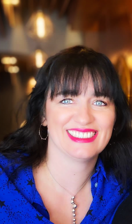 A woman with black hair and blue eyes smiling, wearing a blue shirt with black stars, hoop earrings, and a necklace, in a warmly lit indoor setting.