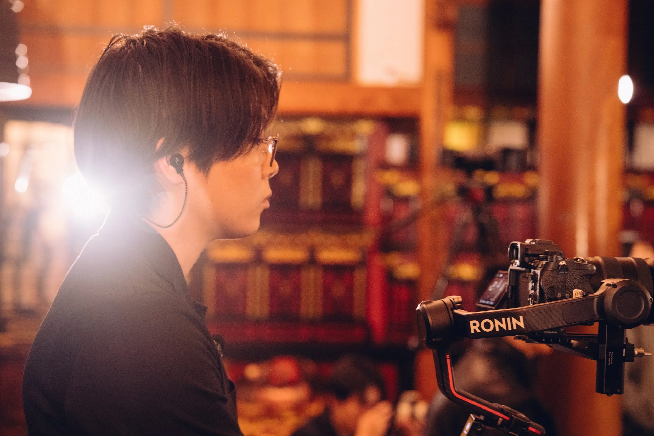 カメラを操作している若い男性が、映画や映像制作の現場で作業している様子。背景は木製の棚や装飾が見える。