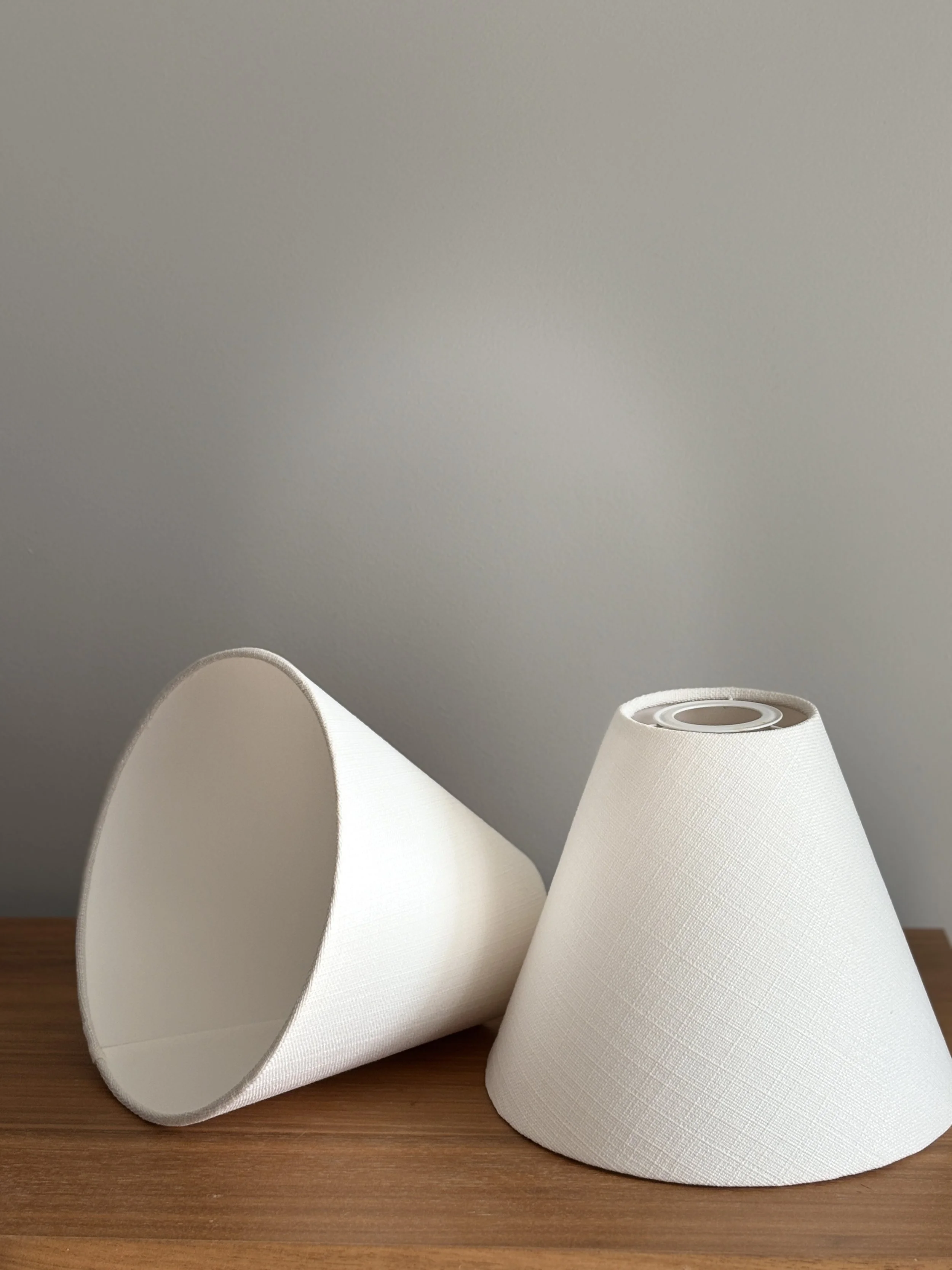 Two white lampshades, one placed upside down and the other upright, on a wooden surface against a plain wall.