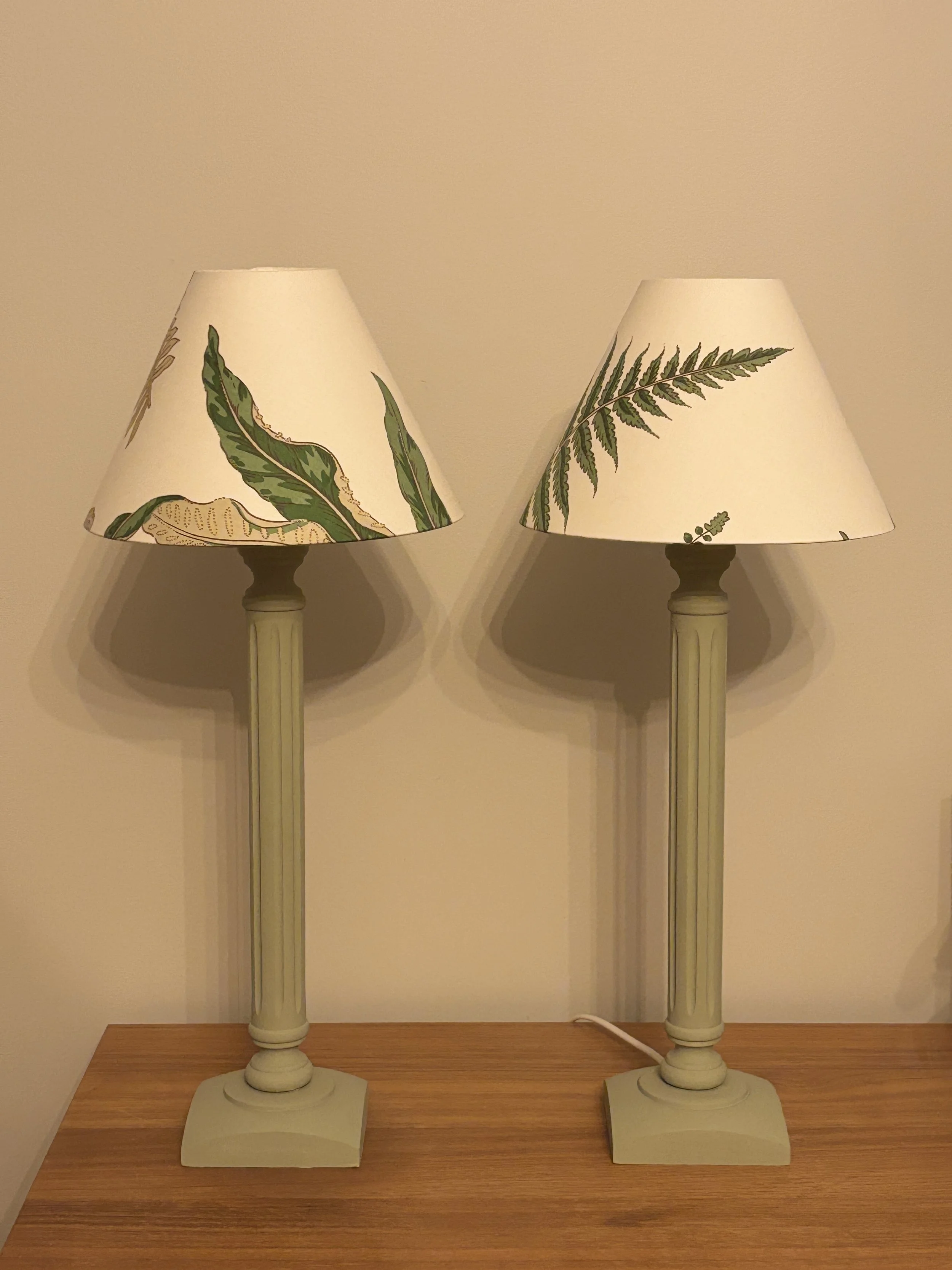 Two table lamps with cream bases and botanical-patterned shades, placed on a wooden surface against a beige wall.