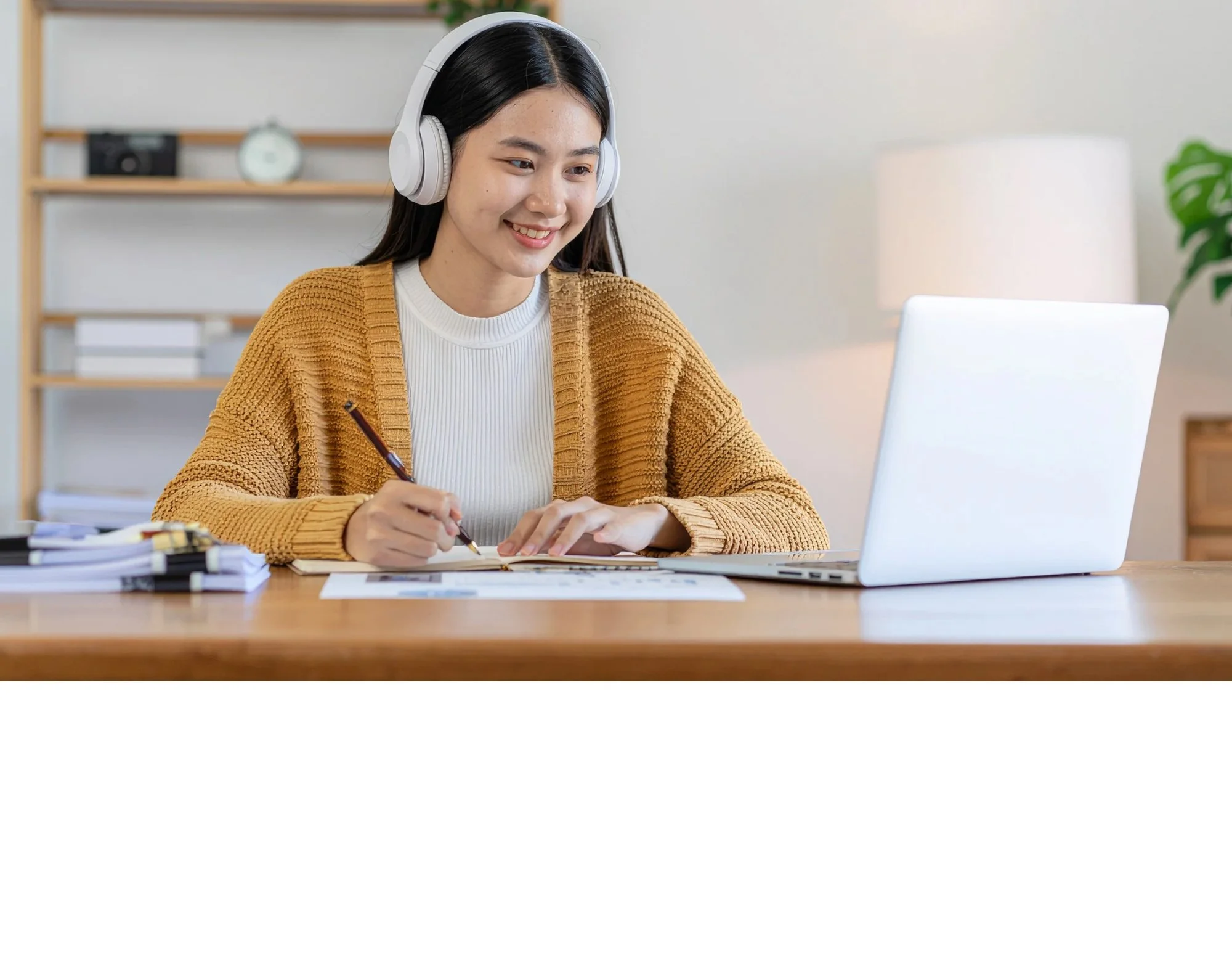 A young woman with long dark hair, wearing a white shirt and a mustard-colored cardigan, sitting at a wooden desk with a silver laptop, headphones, and various papers, smiling while writing in a notebook.