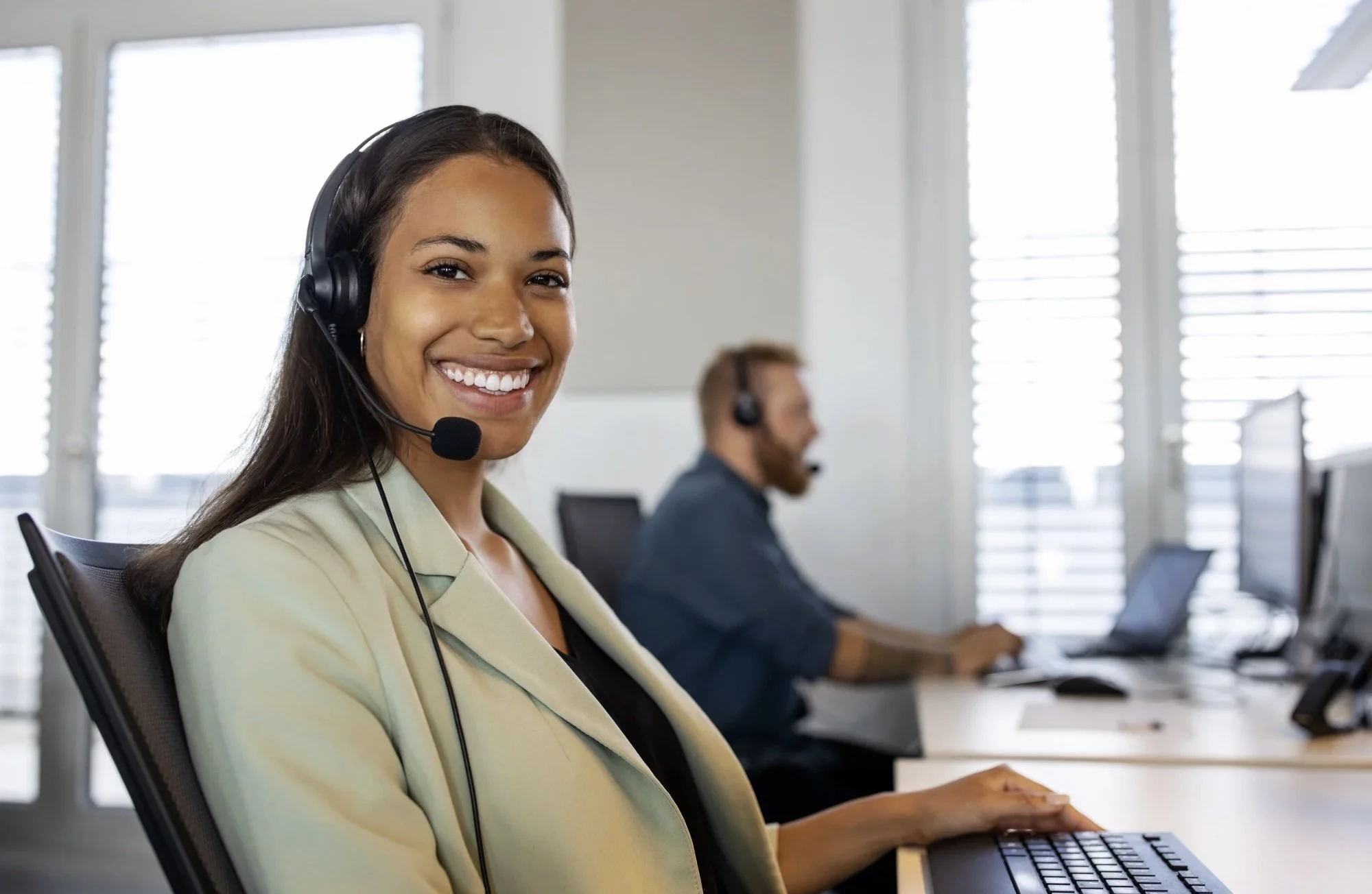 A smiling woman wearing a headset and a beige blazer working at a desk in a bright office with two computer monitors.