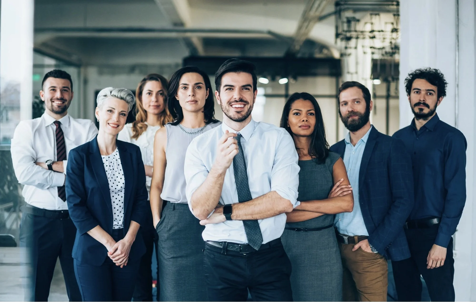 A diverse group of eight professionals standing confidently in a modern office environment, with some smiling and others looking serious, dressed in business casual and formal attire.