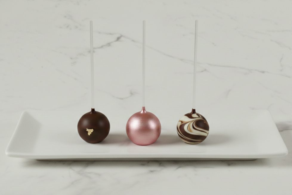 Three decorative cake pops with lollipop sticks on a white rectangular plate, against a white marble background.