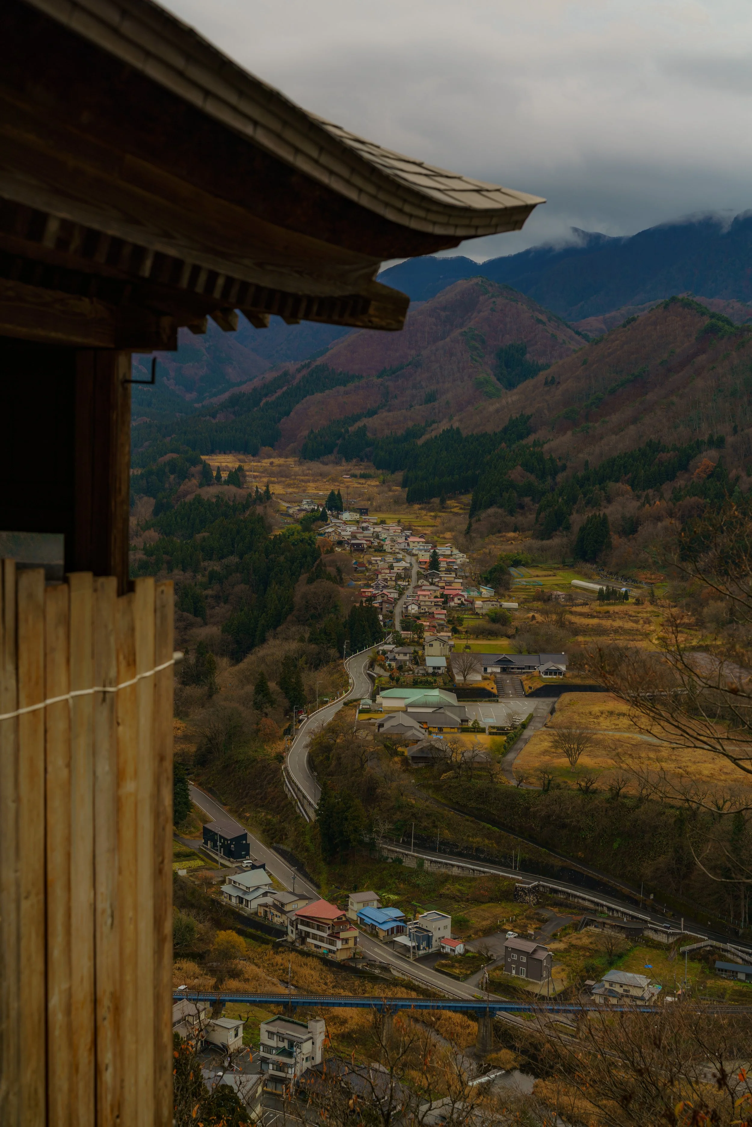 Aerial view of a winding mountain road in a rural area with houses and fields, seen from a balcony of a traditional wooden building on a cloudy day.