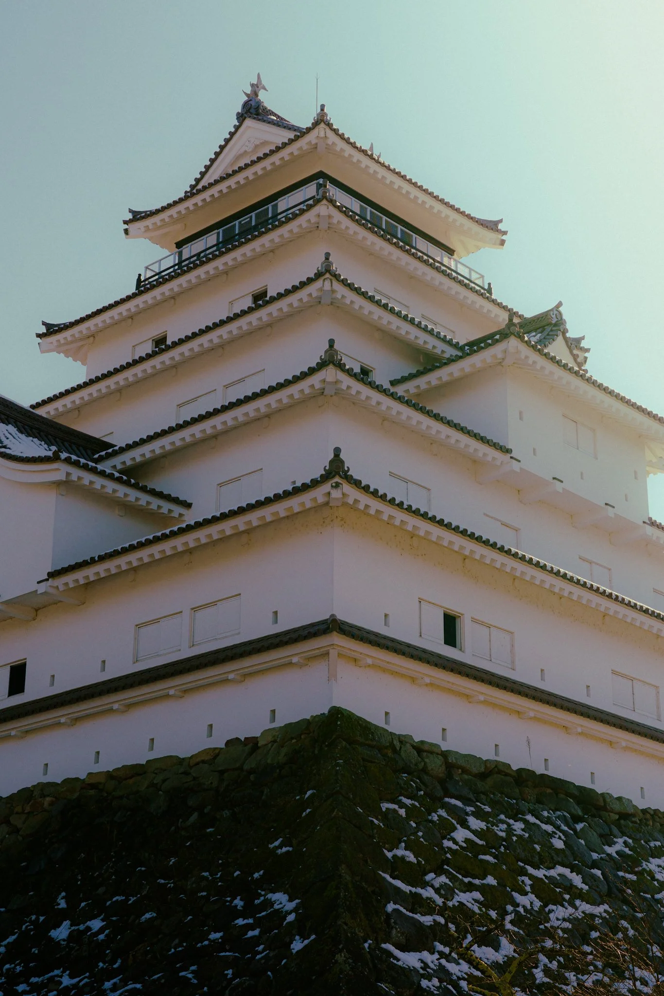 A traditional Japanese castle with white walls and dark tiled roofs, situated on a stone base with some snow, under a clear sky.