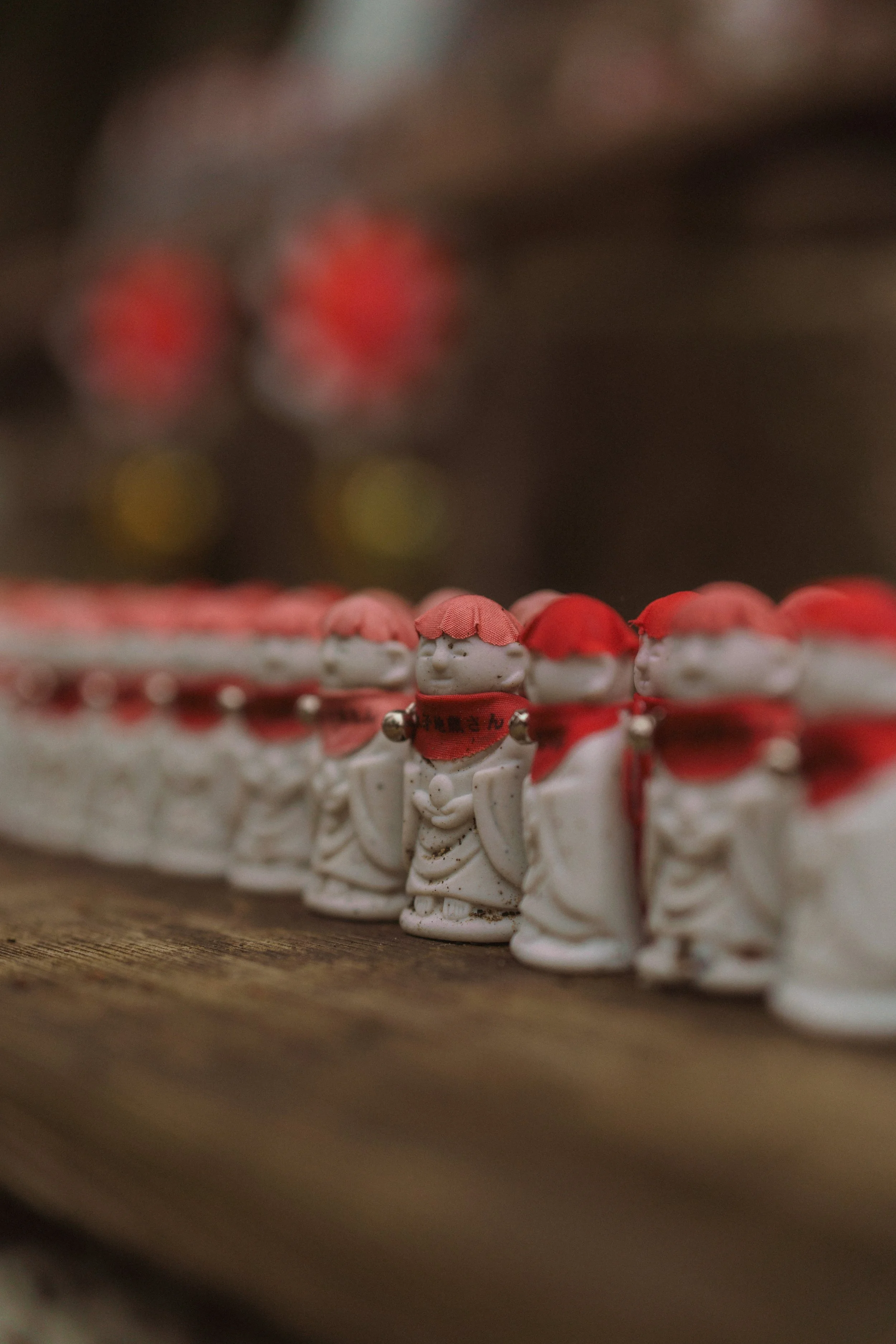 Close-up of small Japanese figurines with pink hats and red scarves, arranged in a row on a wooden surface, with blurred red and yellow decorations in the background.