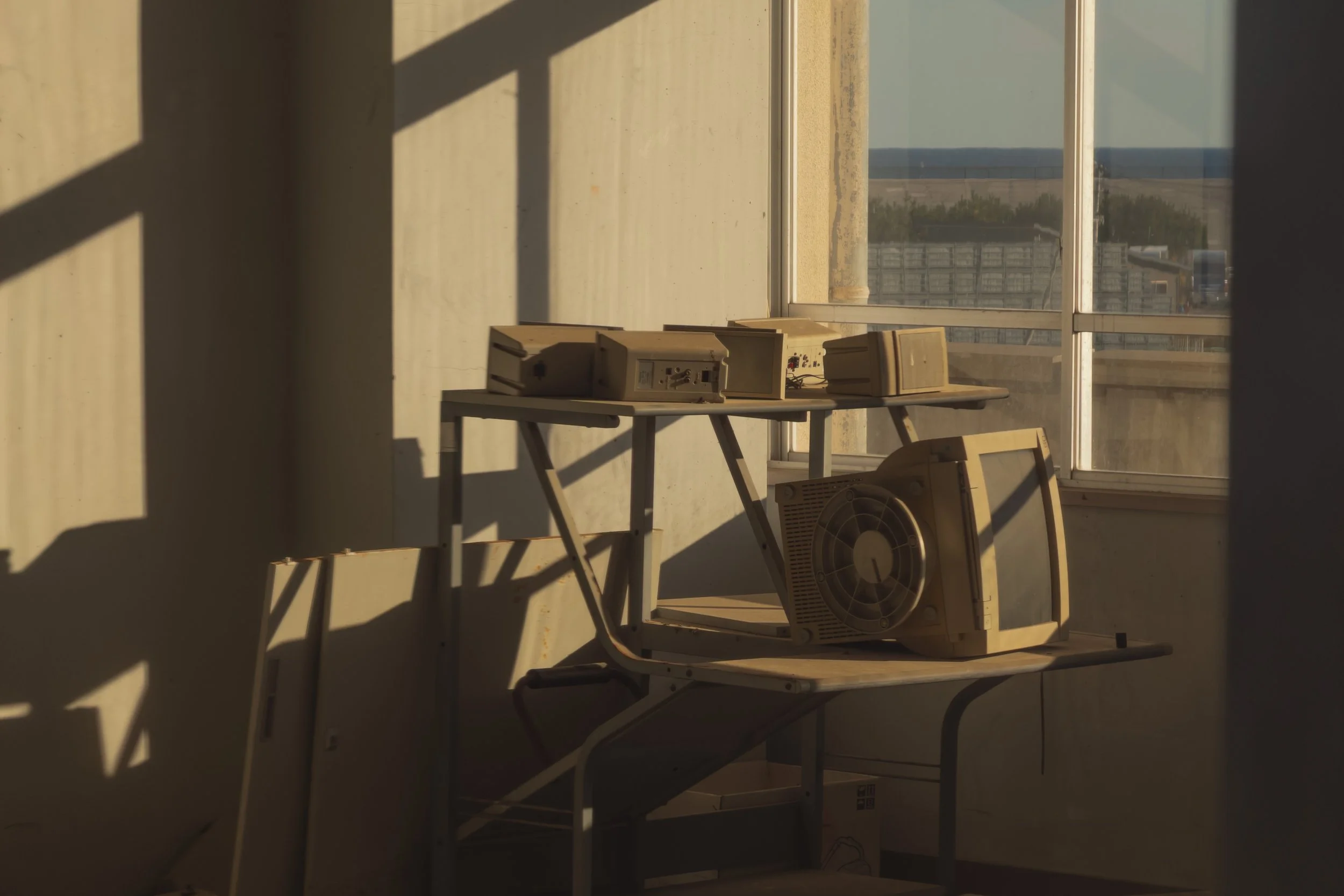 Old beige computer monitors and hardware on a table near large window, sunlight casting shadows inside a room.