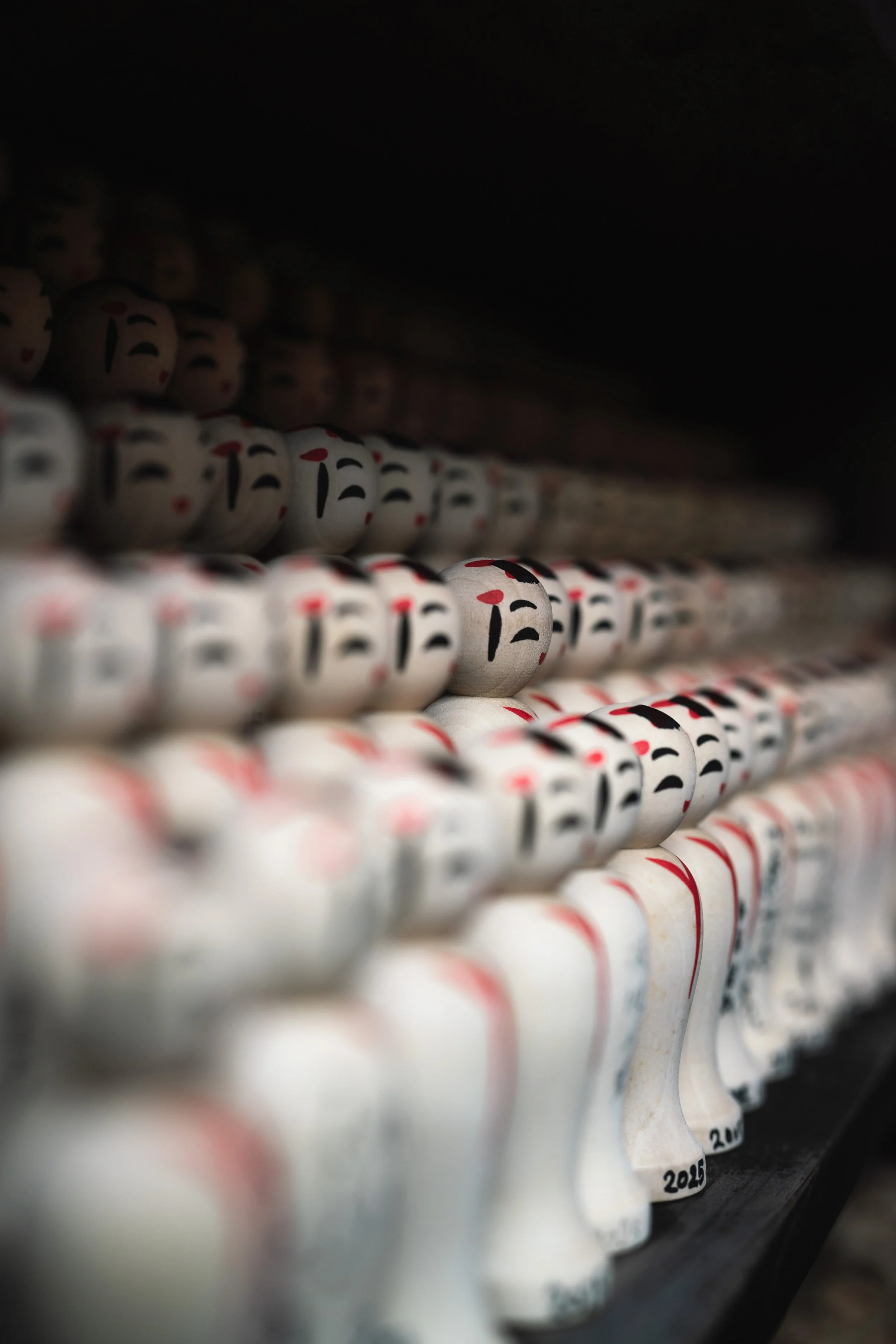 A close-up of multiple white ceramic figurines arranged in rows, each with black and red painted facial features, on a dark shelf.