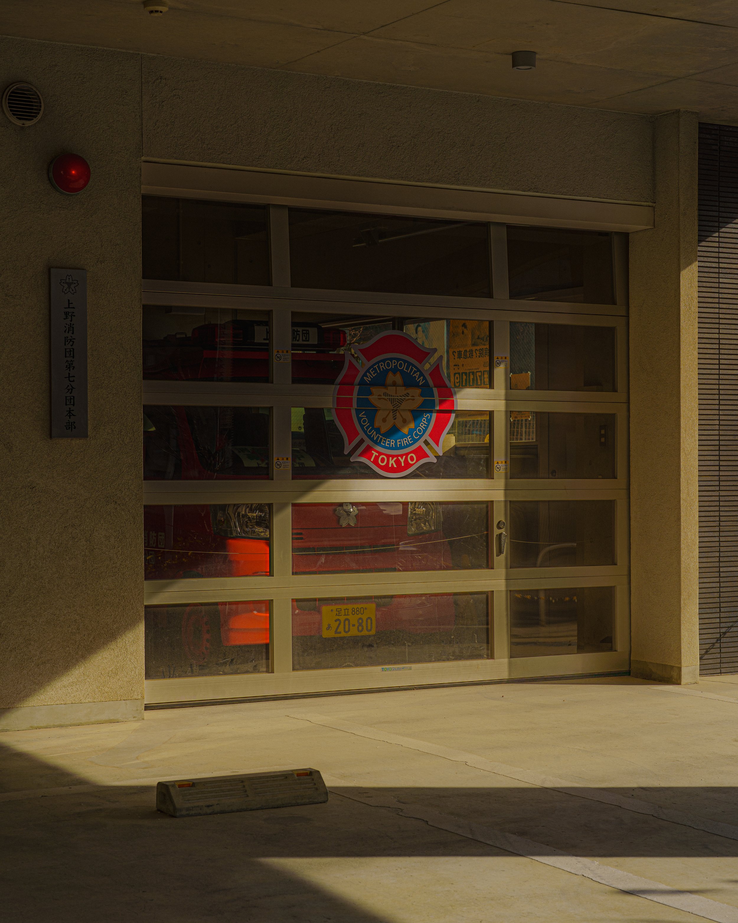 A garage door with a Tokyo Metropolitan Volunteer Fire Corps sign, a fire truck inside, and Japanese license plates, exterior of a building.