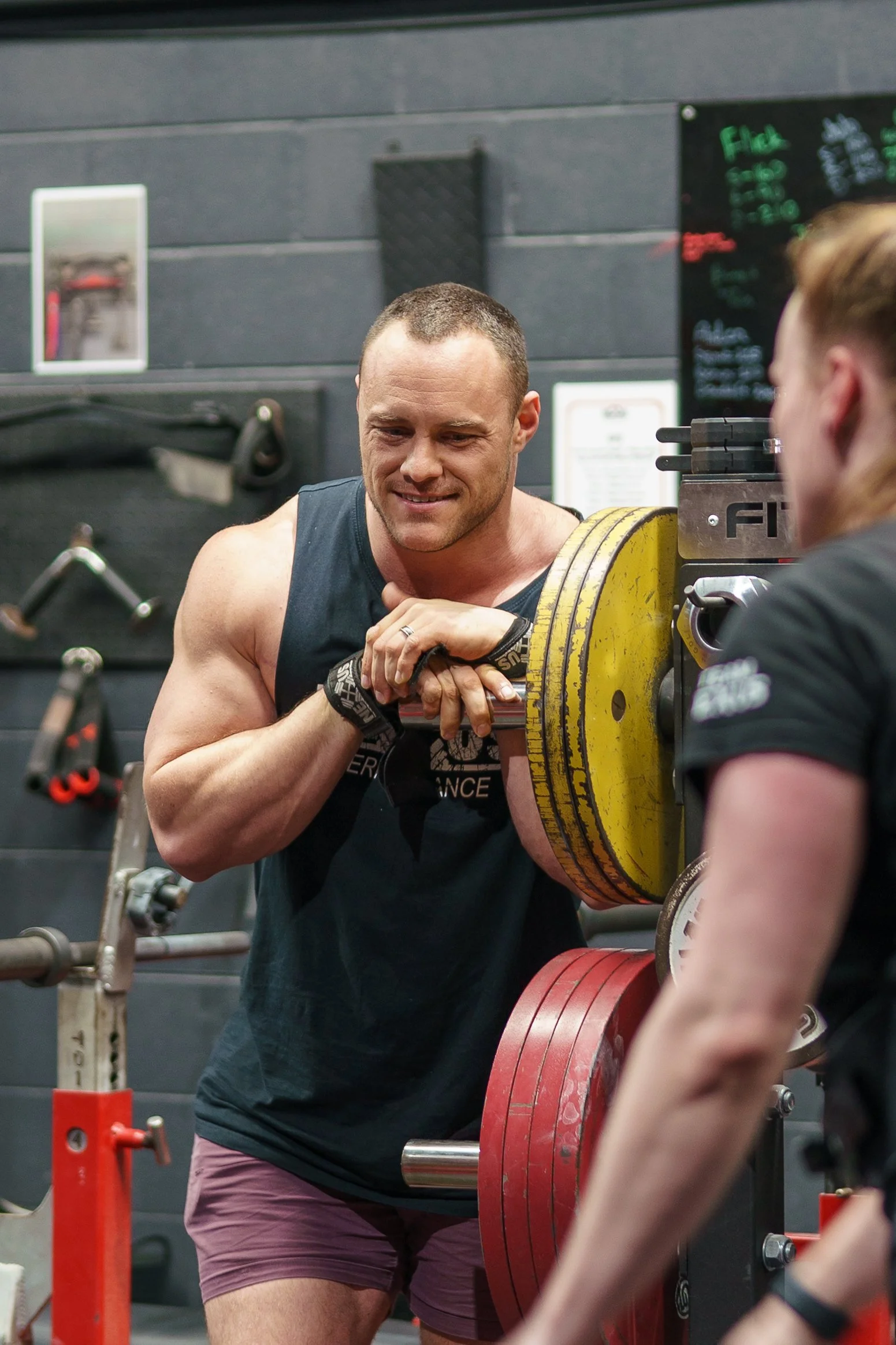 A muscular man smiling, wearing a black tank top, standing in a gym, preparing to lift a barbell with large yellow and red weights, with a woman facing him.