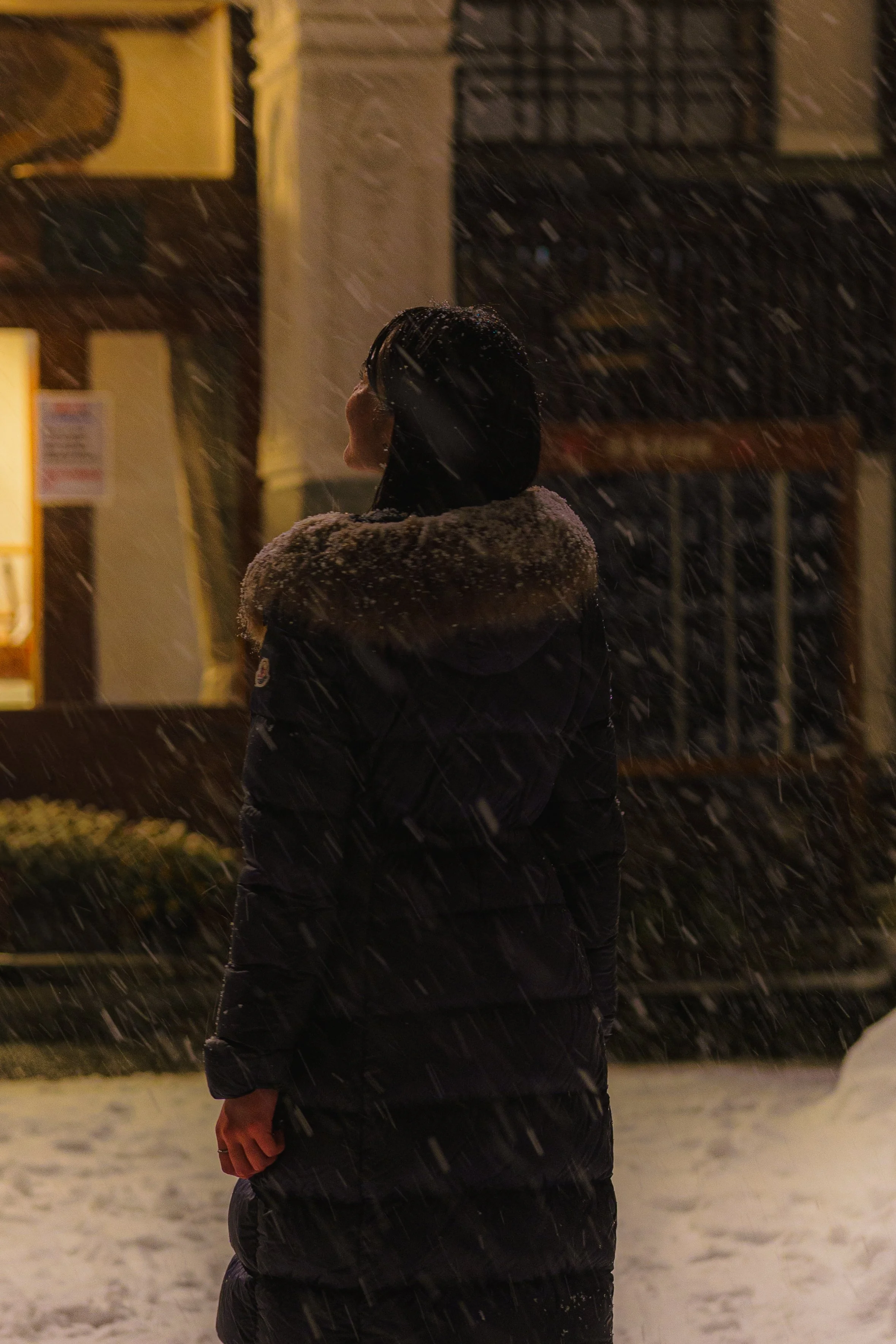 A person standing outside in snowy weather at night, wearing a black winter coat with a fur-lined hood, looking up as snow falls around them.