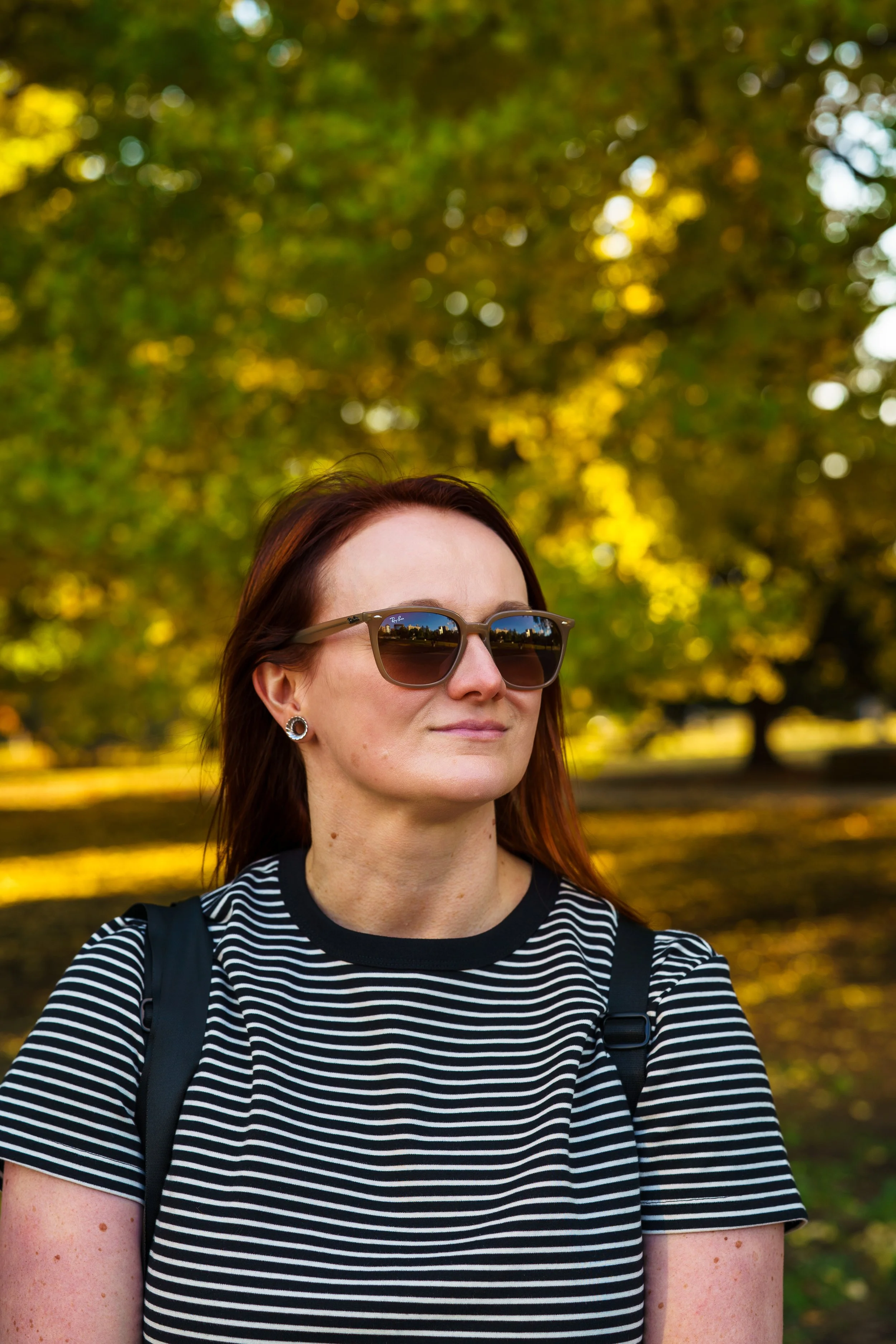 Woman with dark brown hair wearing sunglasses, a black and white striped shirt, and a black backpack outdoors with trees and sunlight in the background.