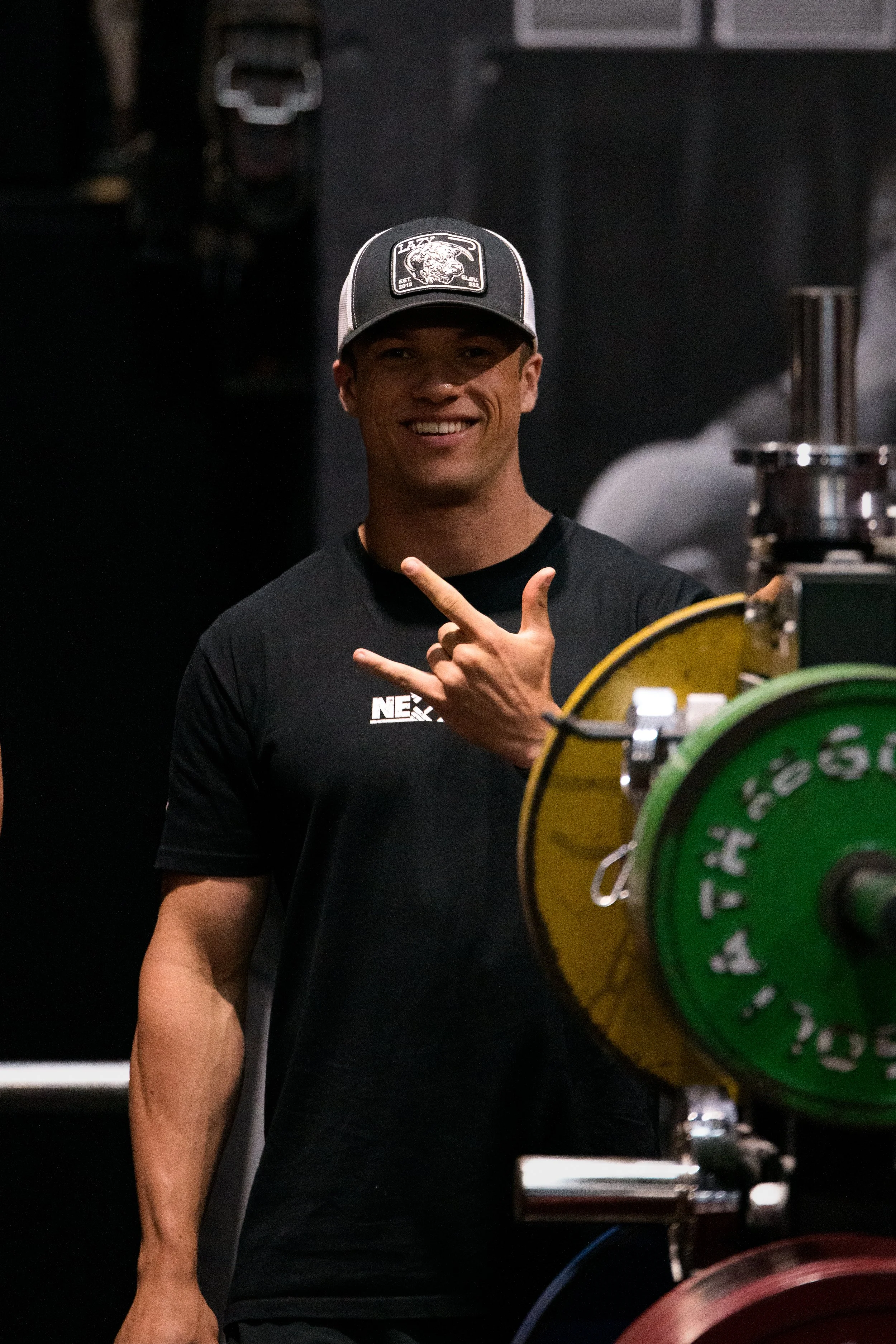 A man smiling and making a hand gesture, standing next to a plate-loaded barbell in a gym.