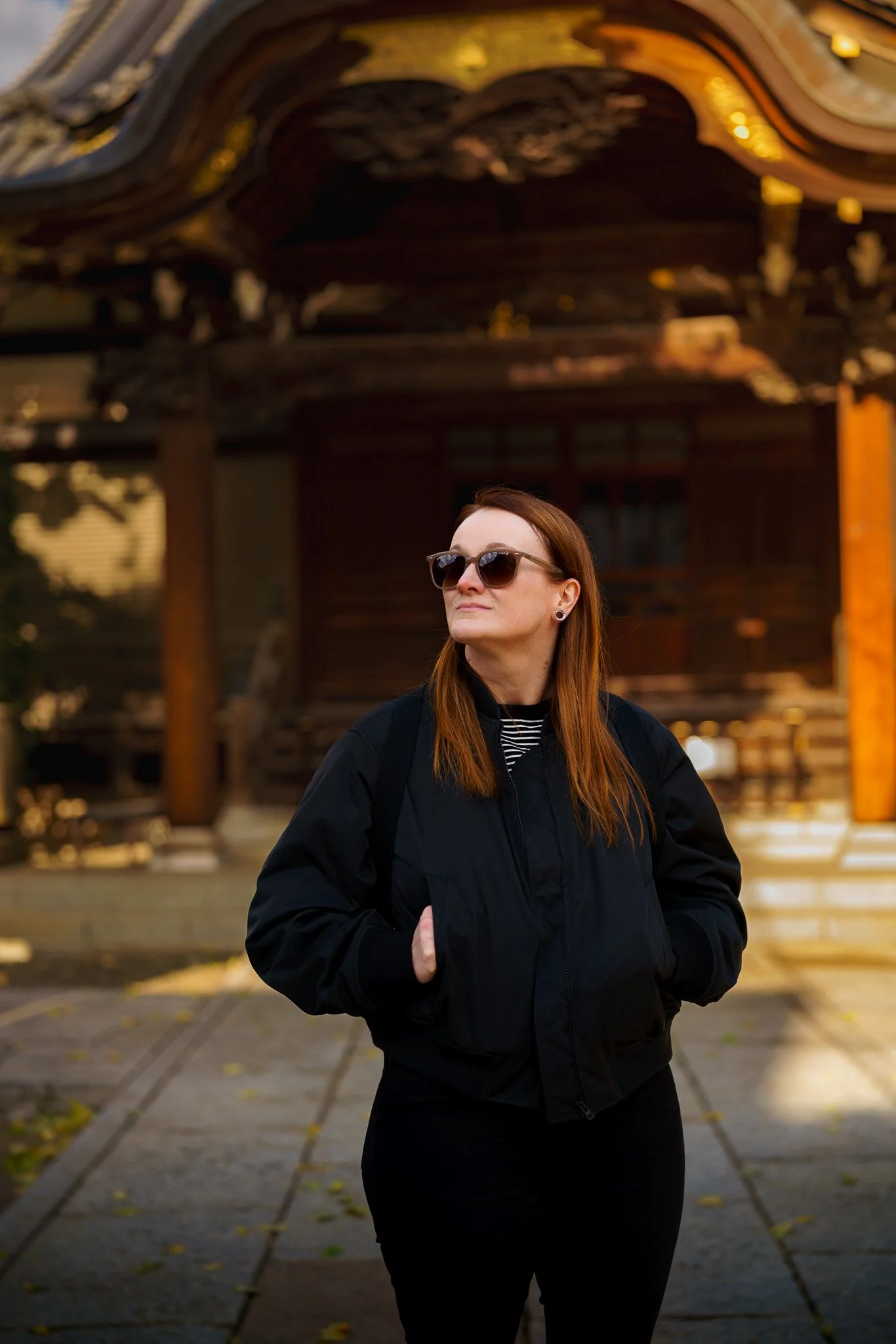 A woman with red hair wearing sunglasses and a black jacket standing outdoors in front of a traditional Japanese shrine.