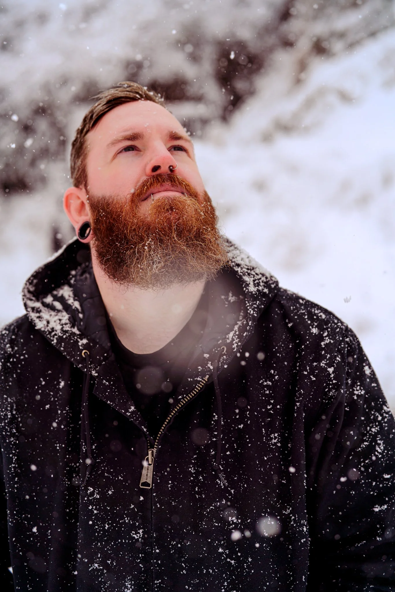 A man with a red beard and piercings looking up in snowy weather, wearing a black hoodie covered in snow.