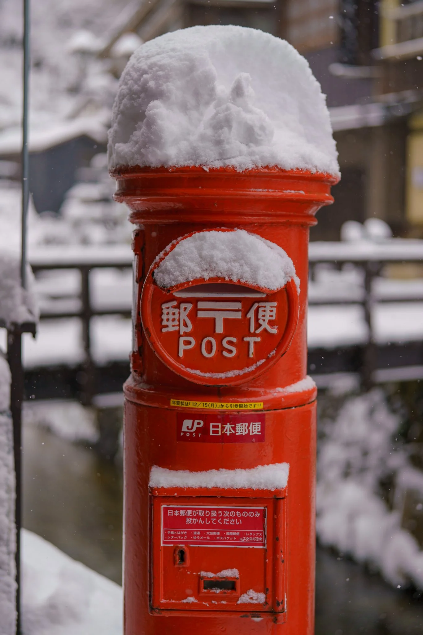 A red Japanese postal mailbox covered in snow, with snow on top and on the front, displaying the postal service logo and Japanese characters.