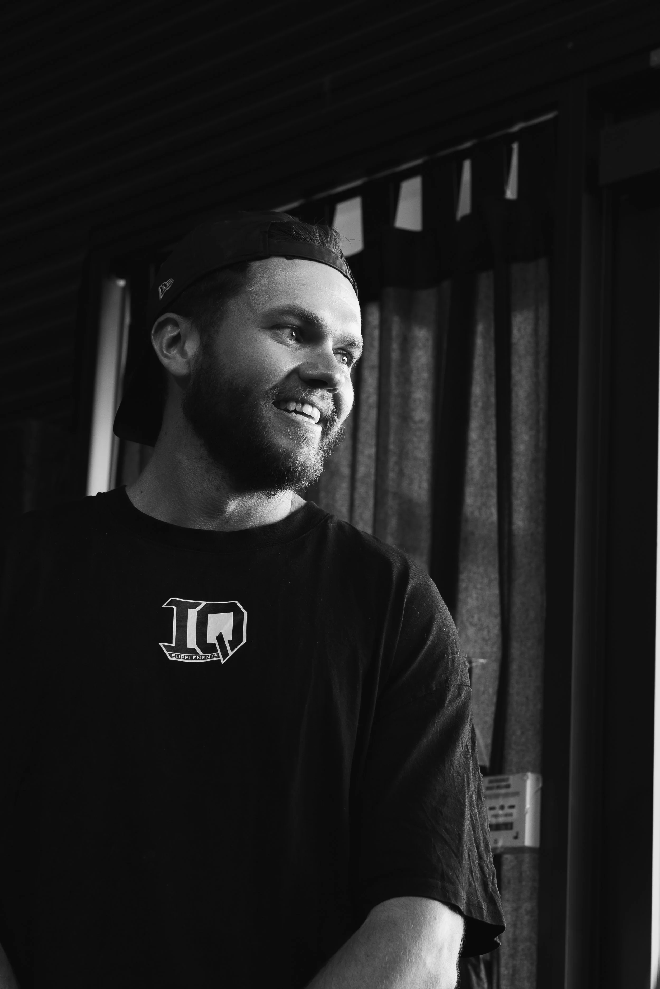 A young man with a beard and a backward cap smiling, indoors near a window with curtains.