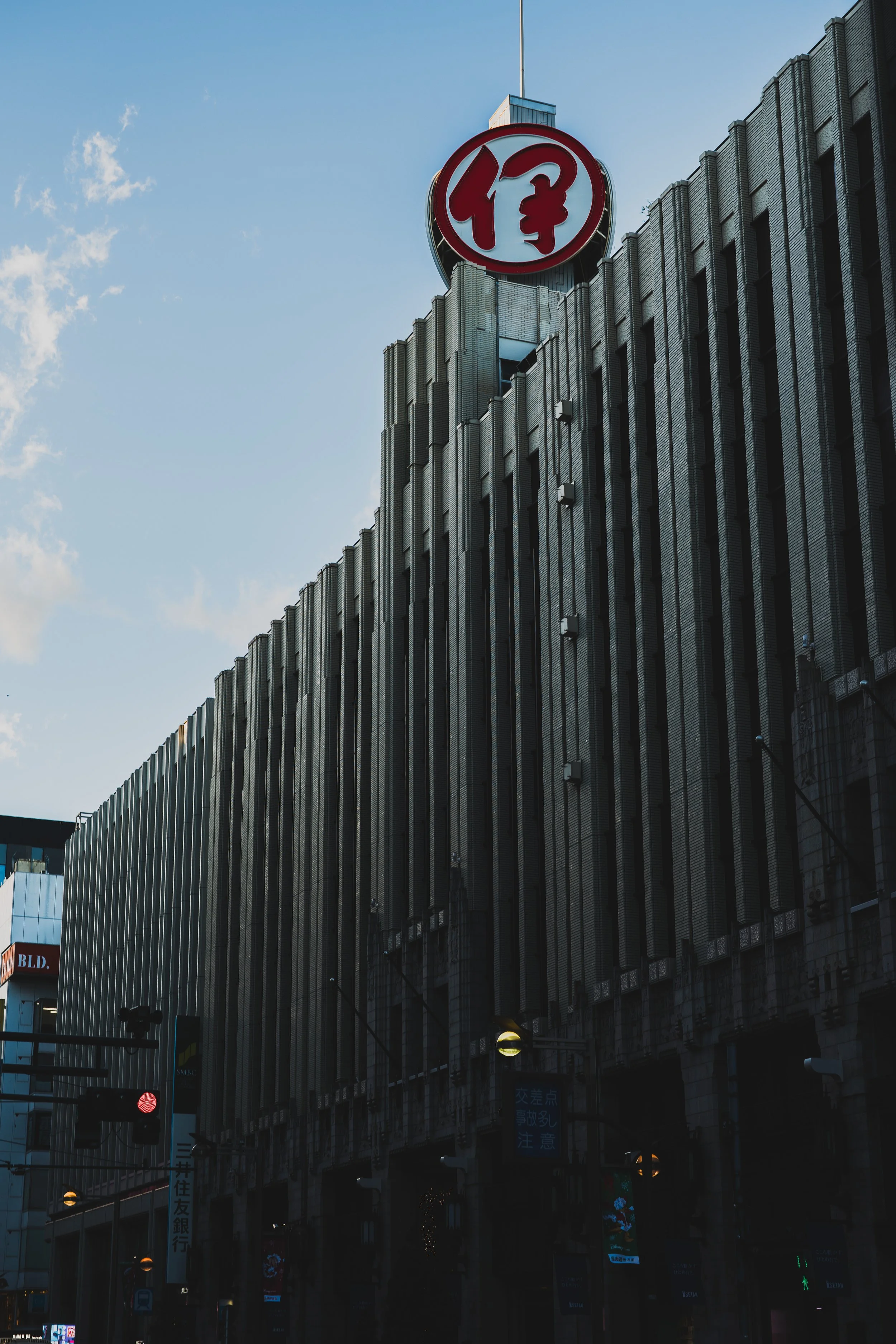 A tall city building with vertical lines and a red circular sign with a white symbol on top. Traffic lights and street signs are visible at street level.