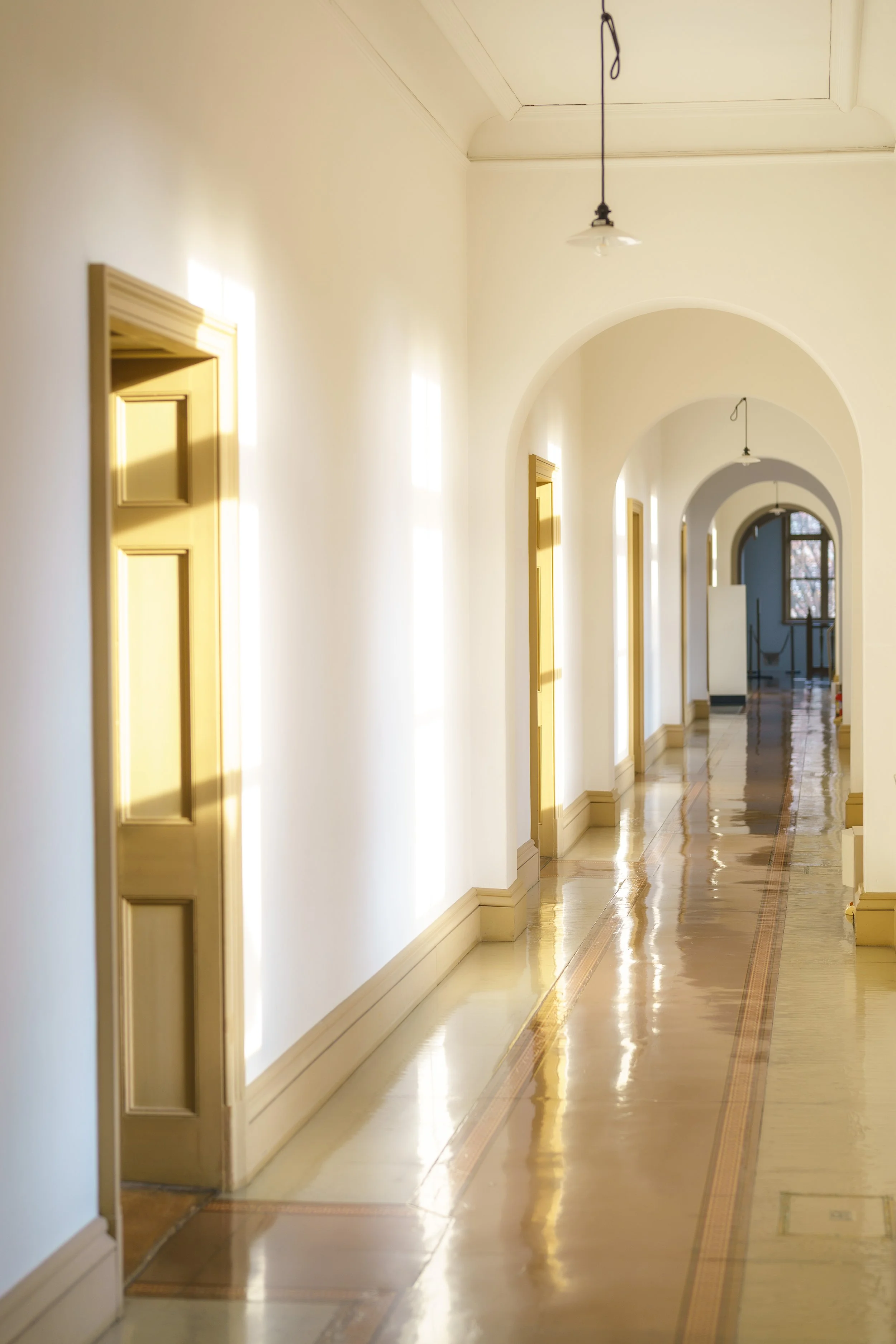 Bright corridor with white walls, arched doorways, and gold-colored doors, illuminated by sunlight and hanging light fixtures.