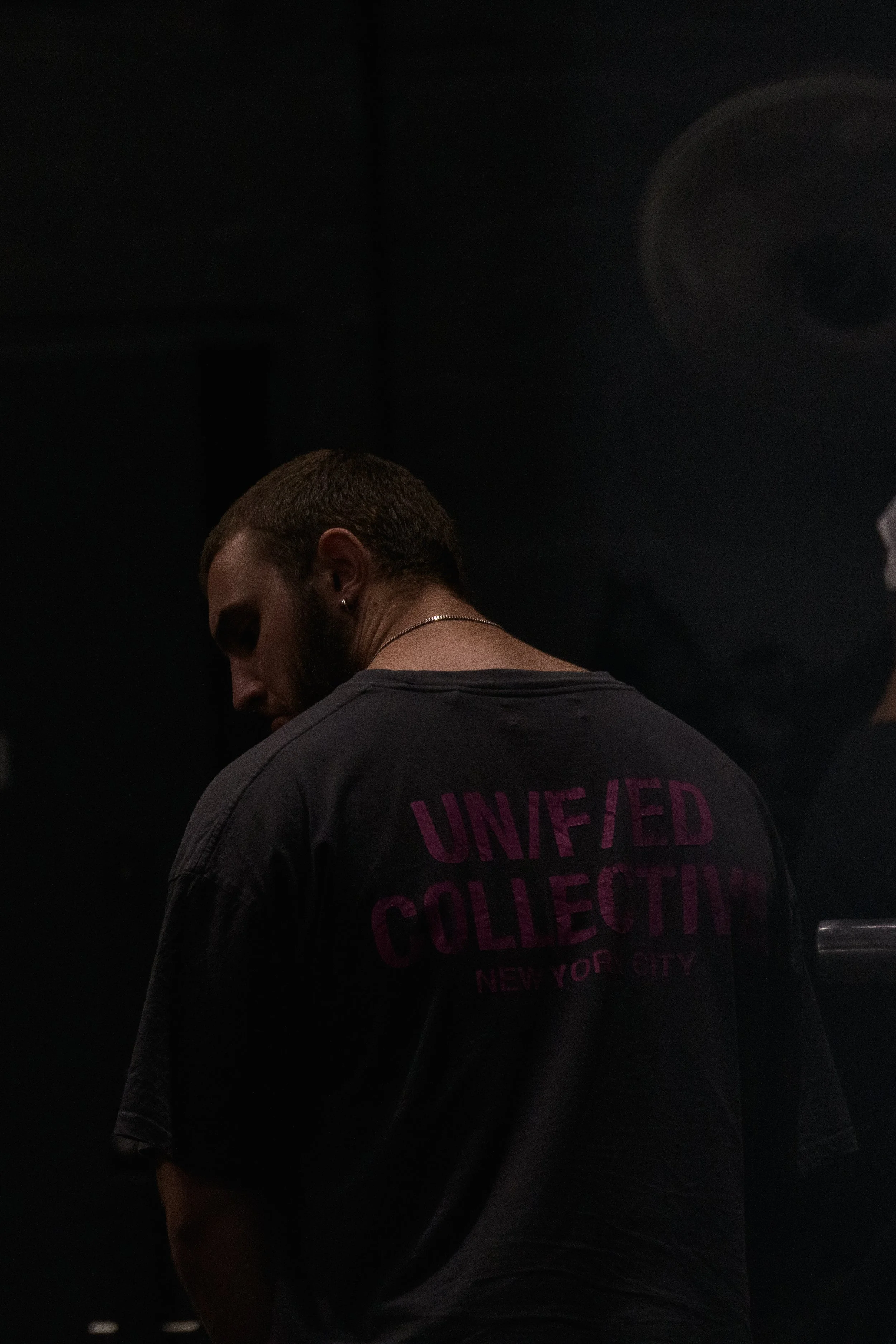 A man with a beard wearing a black T-shirt with magenta text that says 'UNIFIED COLLECTIVE, NEW YORK CITY' on the back, standing in a dark room with dim lighting.