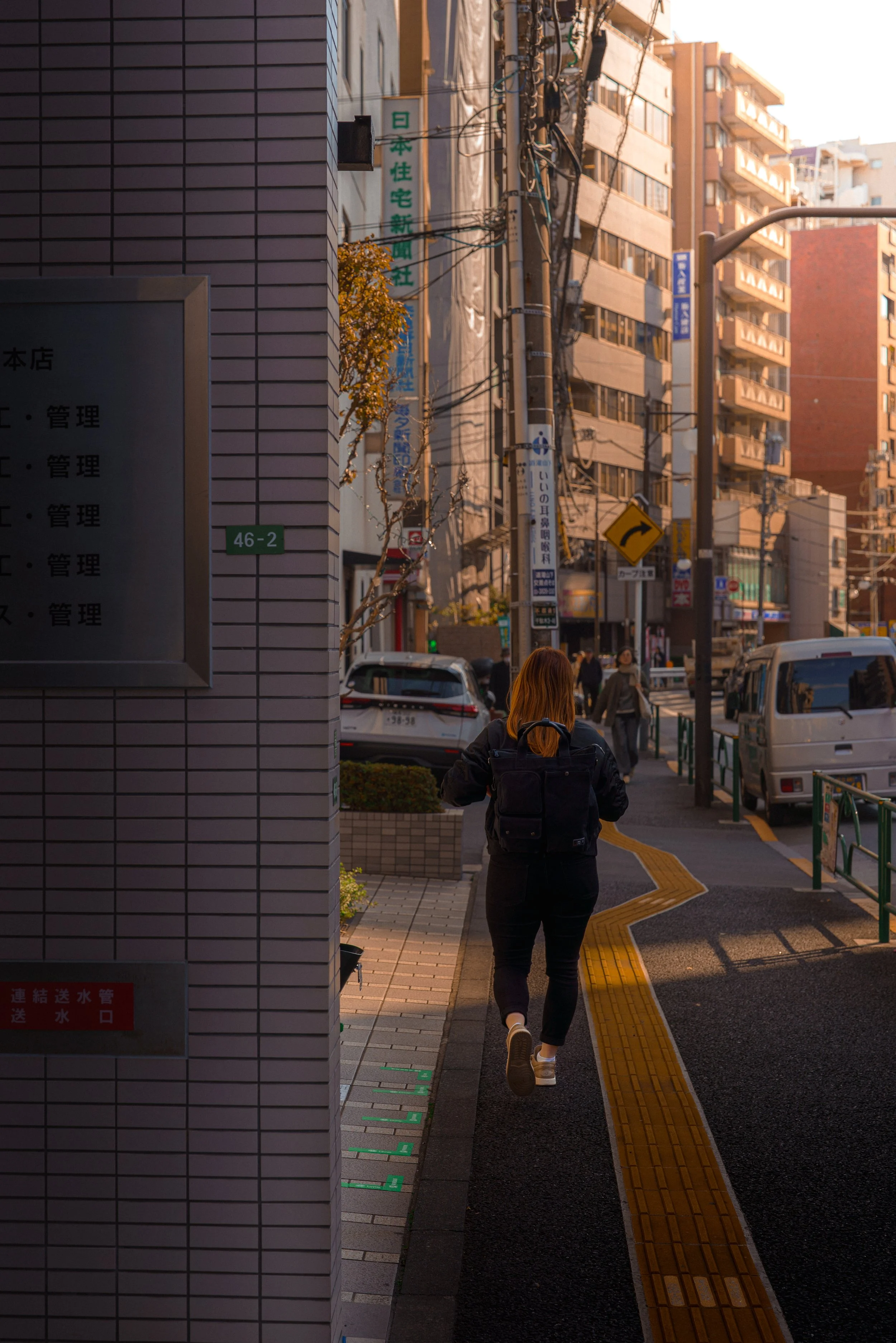 A woman with red hair wearing a black jacket, black pants, and white shoes walking down a sidewalk in an urban area with high-rise buildings, street signs, and cars parked nearby.