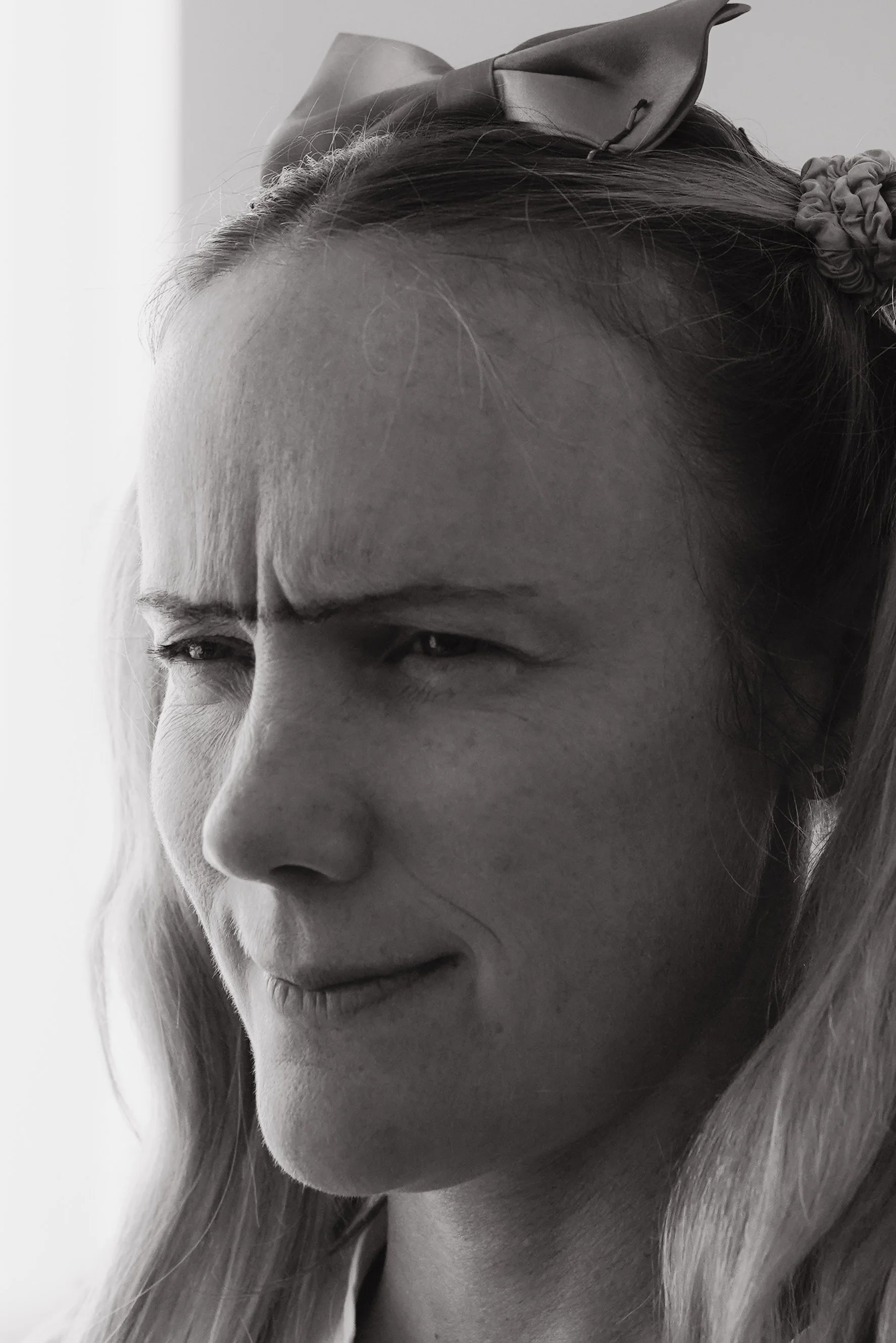 Black and white close-up of a woman with light-colored hair, wearing a bow headband, squinting and showing a slight smirk.