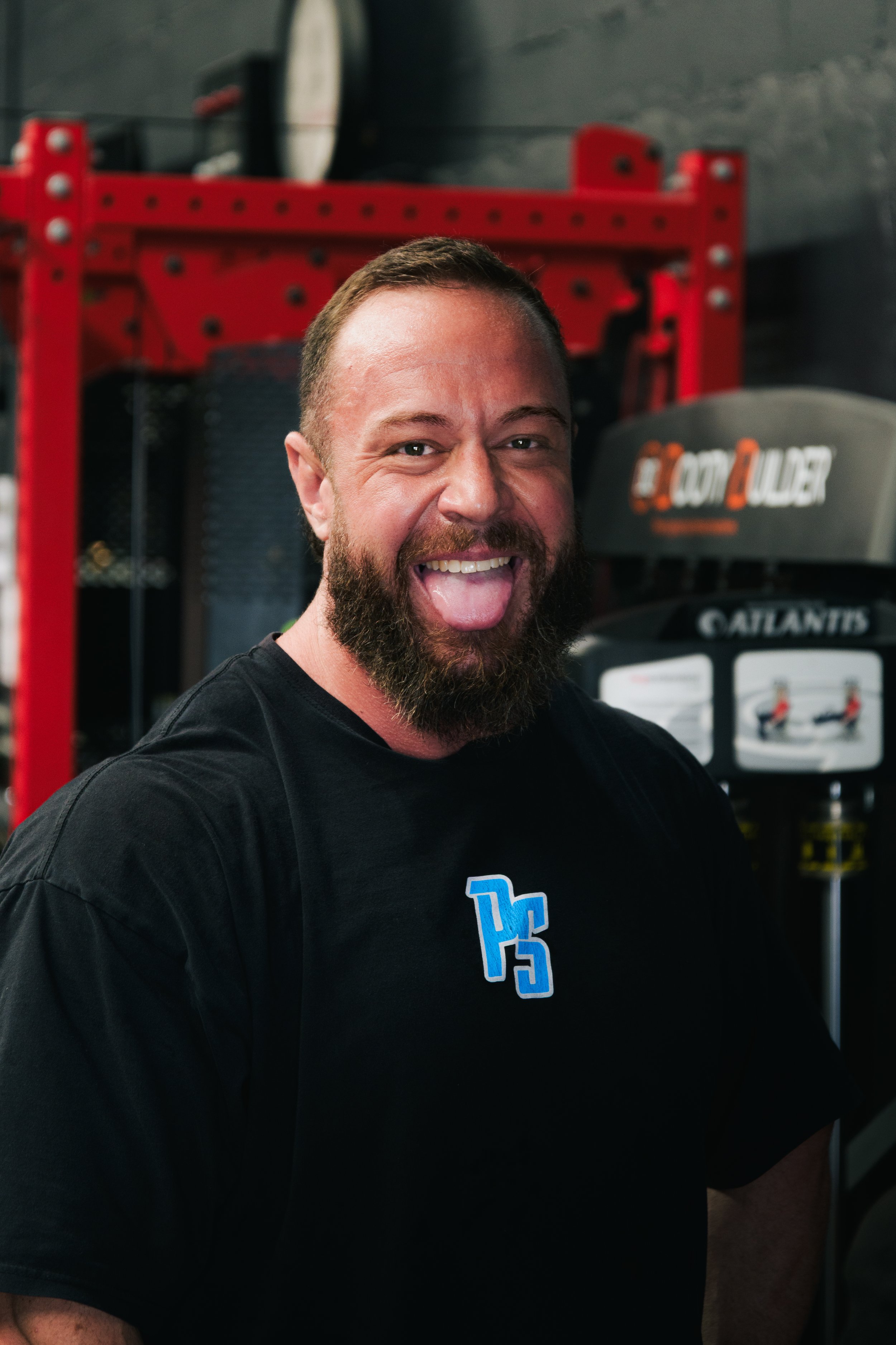 A smiling man with a beard playfully sticking out his tongue at the gym, standing in front of workout equipment.