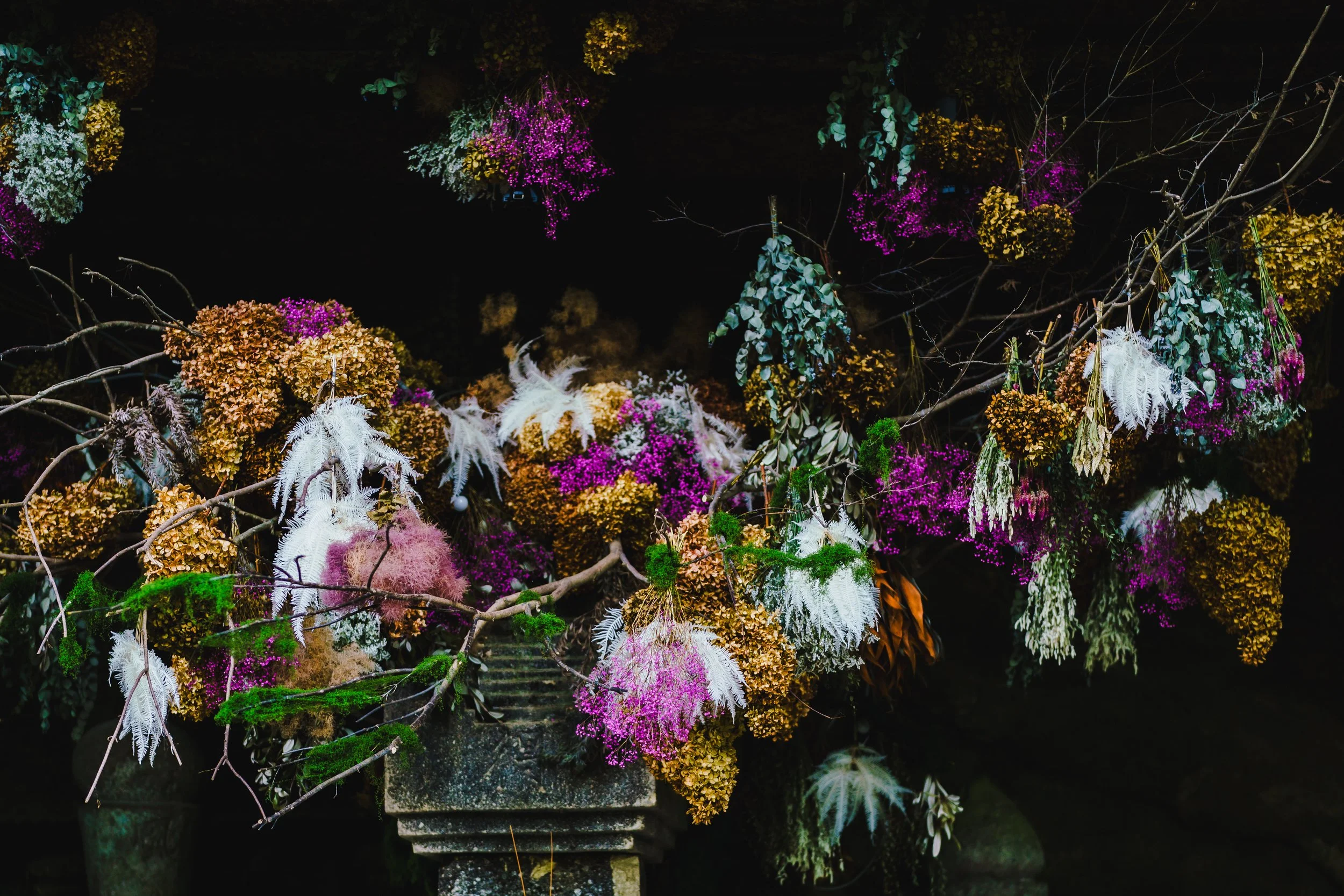 A vibrant display of autumnal flowers and dried plants arranged on a stone pedestal, including colorful hydrangeas, feathers, moss, and twigs against a dark background.