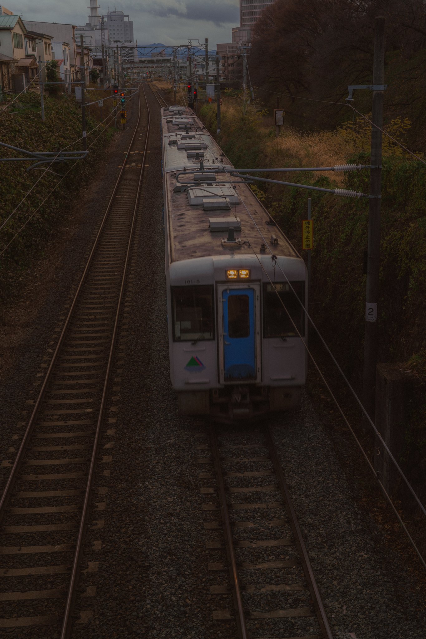A train traveling on railway tracks through an urban area with buildings and trees on either side, under cloudy skies.