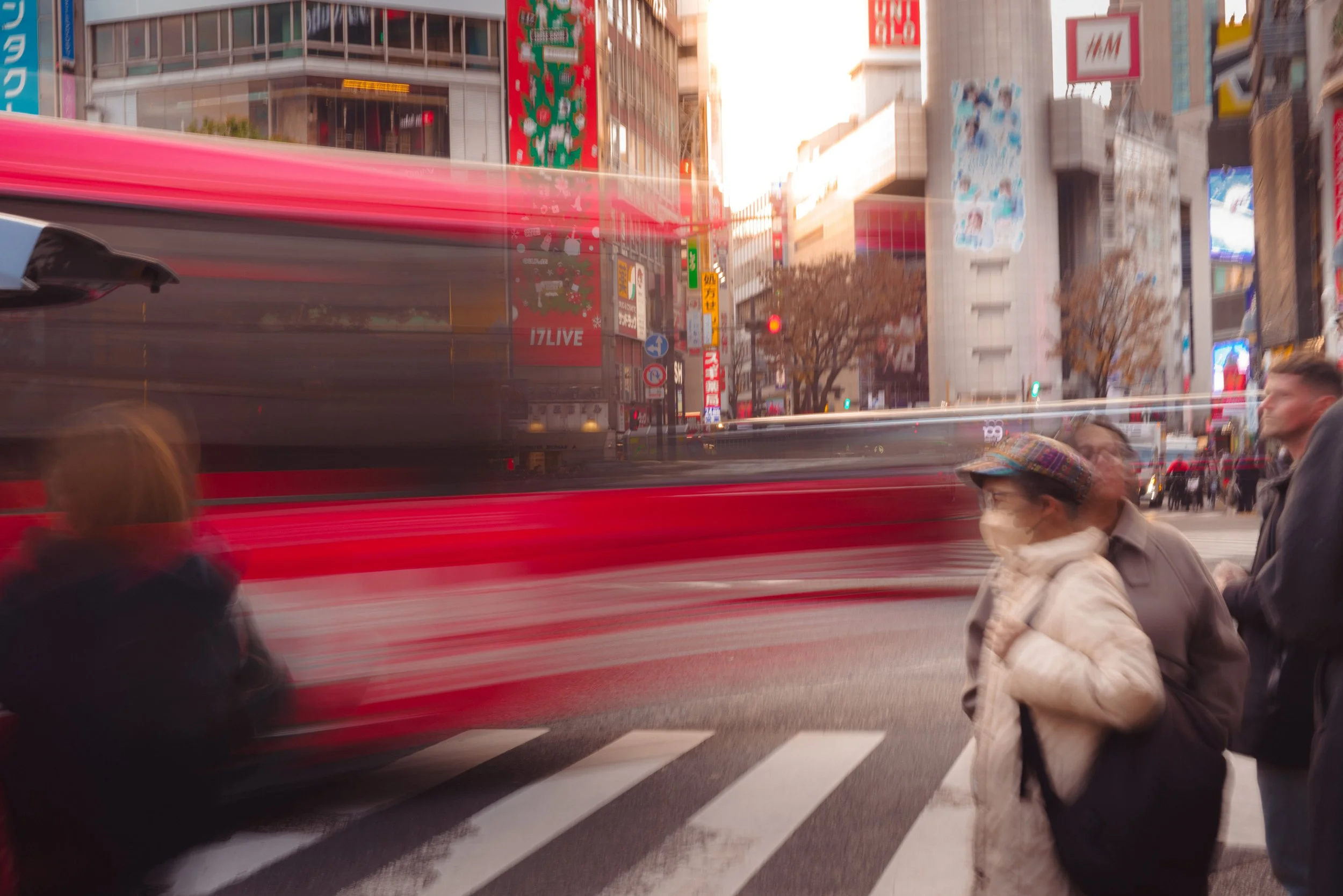 Shibuya crossing at dusk with pedestrians, a blurred red bus moving past, and illuminated billboards and signs in the background.