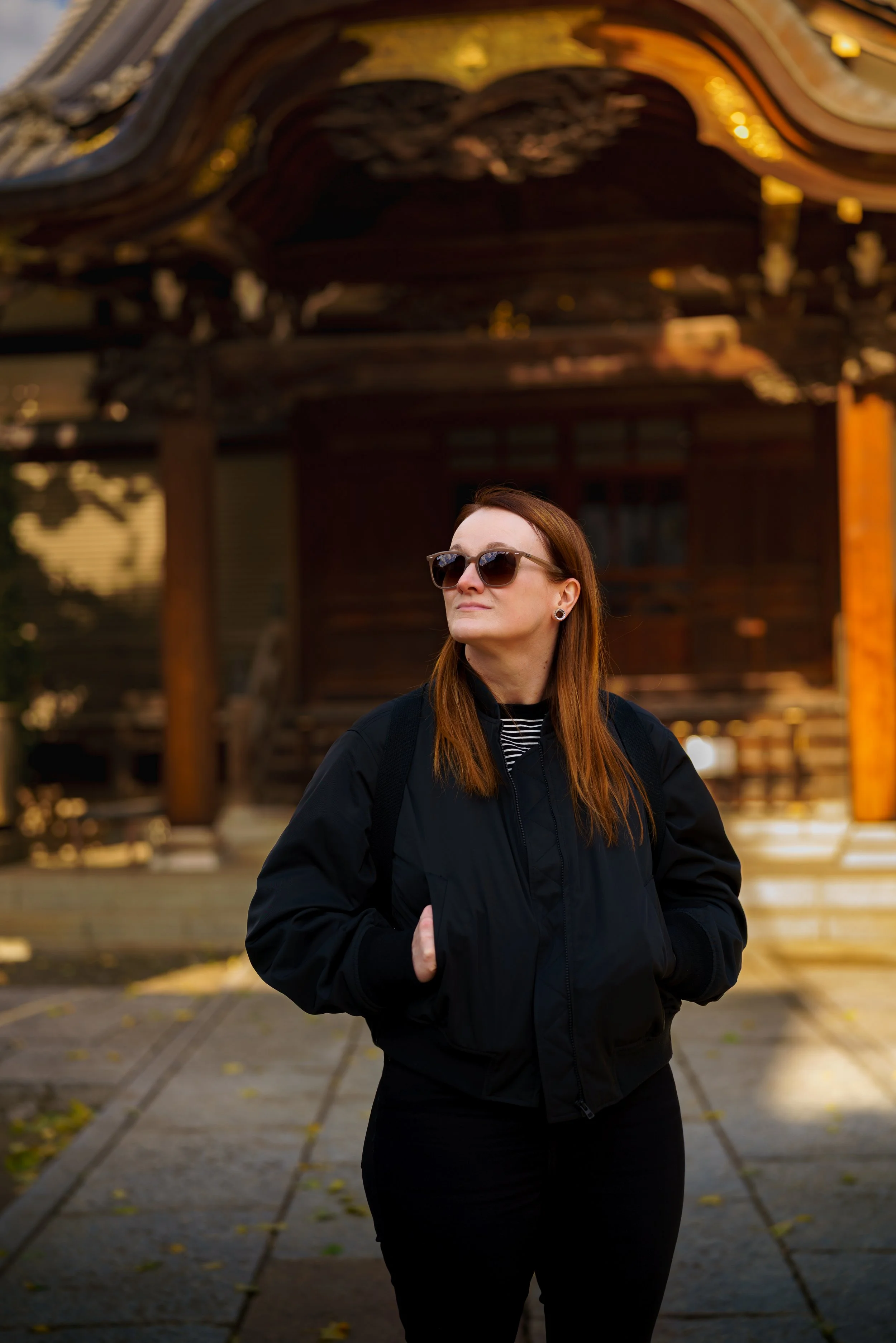 A woman with red hair and sunglasses standing outdoors near a traditional wooden structure, wearing a black jacket and looking off into the distance.