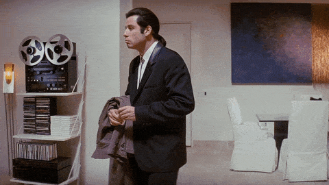 Confused man in a suit holding a coat and looking around a modern living room with a shelf of audio equipment and a painting on the wall.