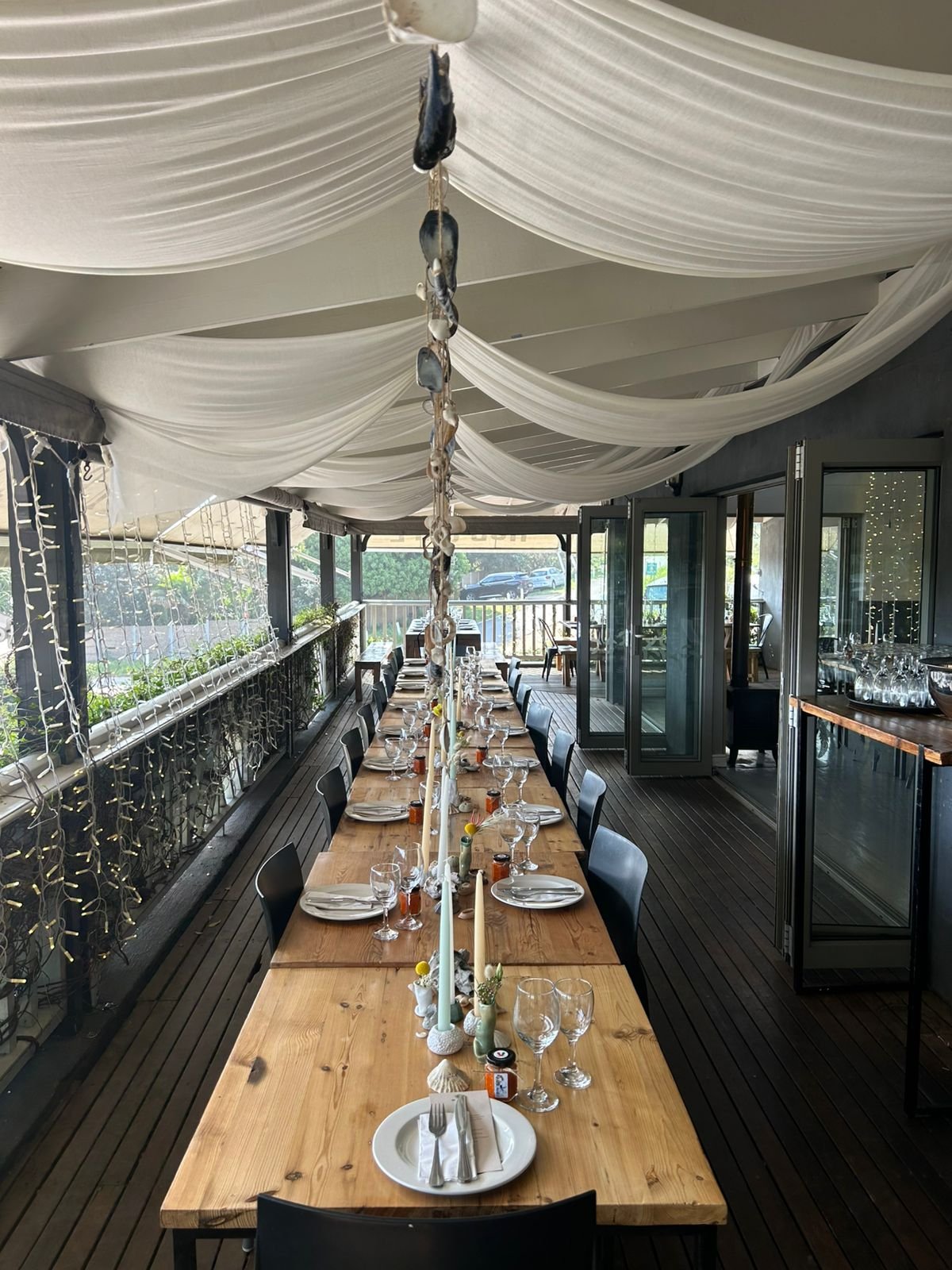Long wooden table set for a meal with plates, glasses, utensils, and candles, in a covered outdoor dining area with white draped fabric ceiling and string lights, overlooking a patio and trees.