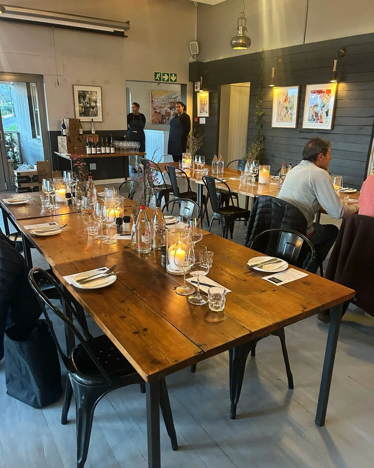 A dining area in a restaurant with a long wooden table set with plates, glasses, and candles, and some people seated and standing in the background.