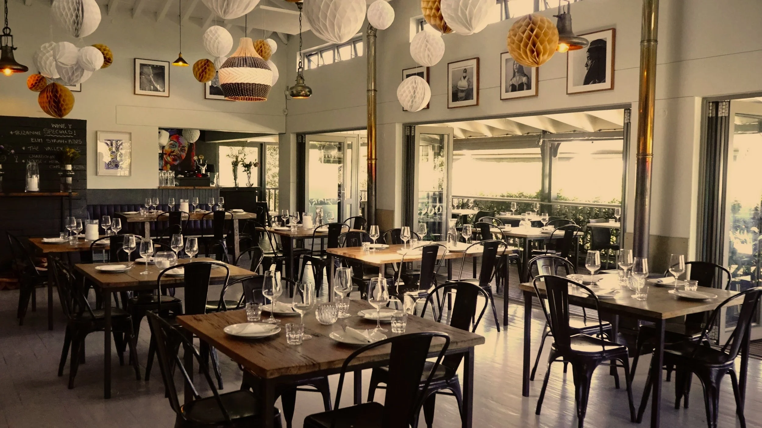 Empty restaurant dining area with wooden tables, black chairs, and wine glasses set for service. Bright natural light from large windows, hanging paper lanterns, photographs on the walls, and a visible outdoor patio.