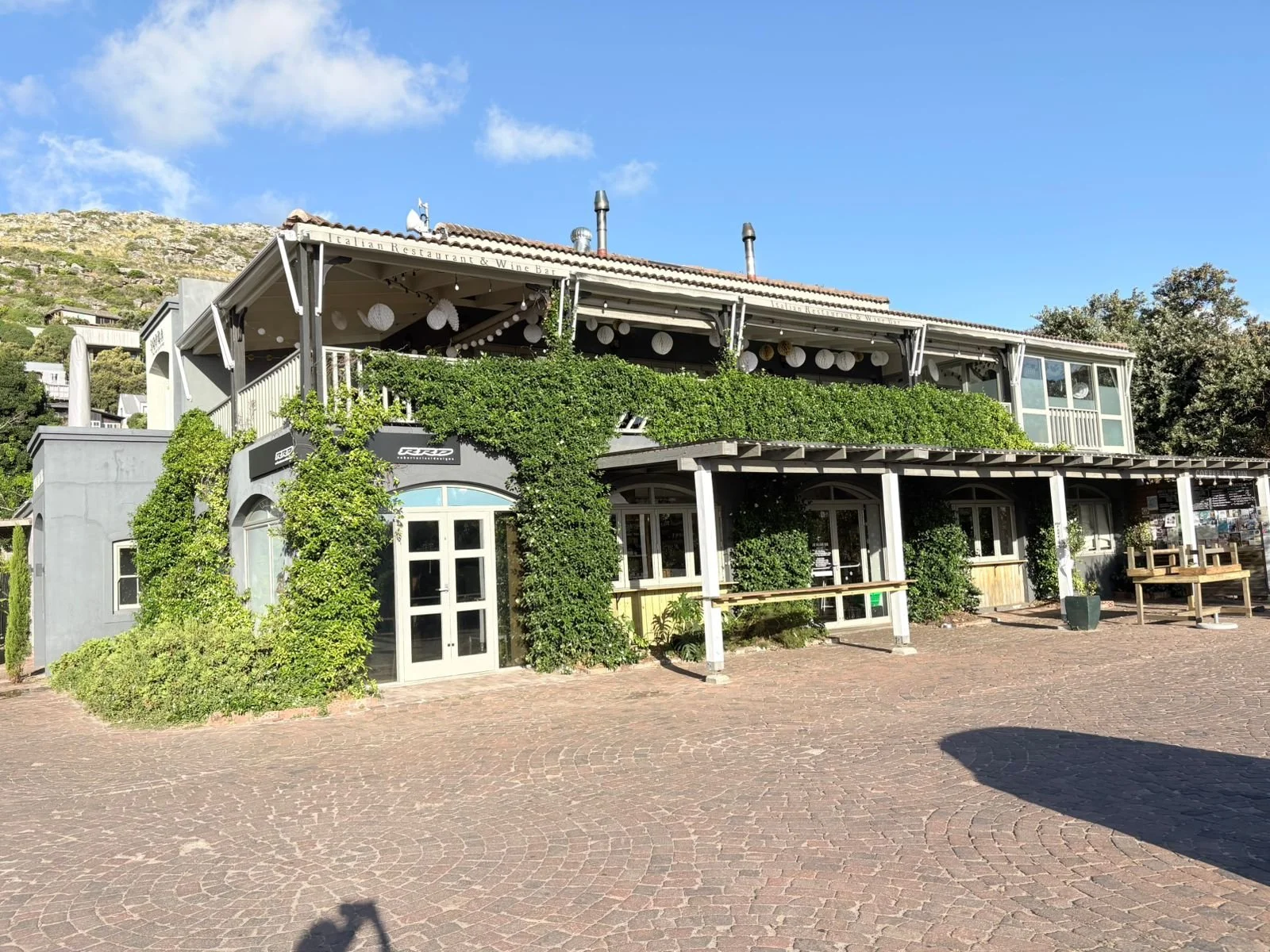 Two-story restaurant building with outdoor seating, green plants, and string lights on a sunny day with blue sky.