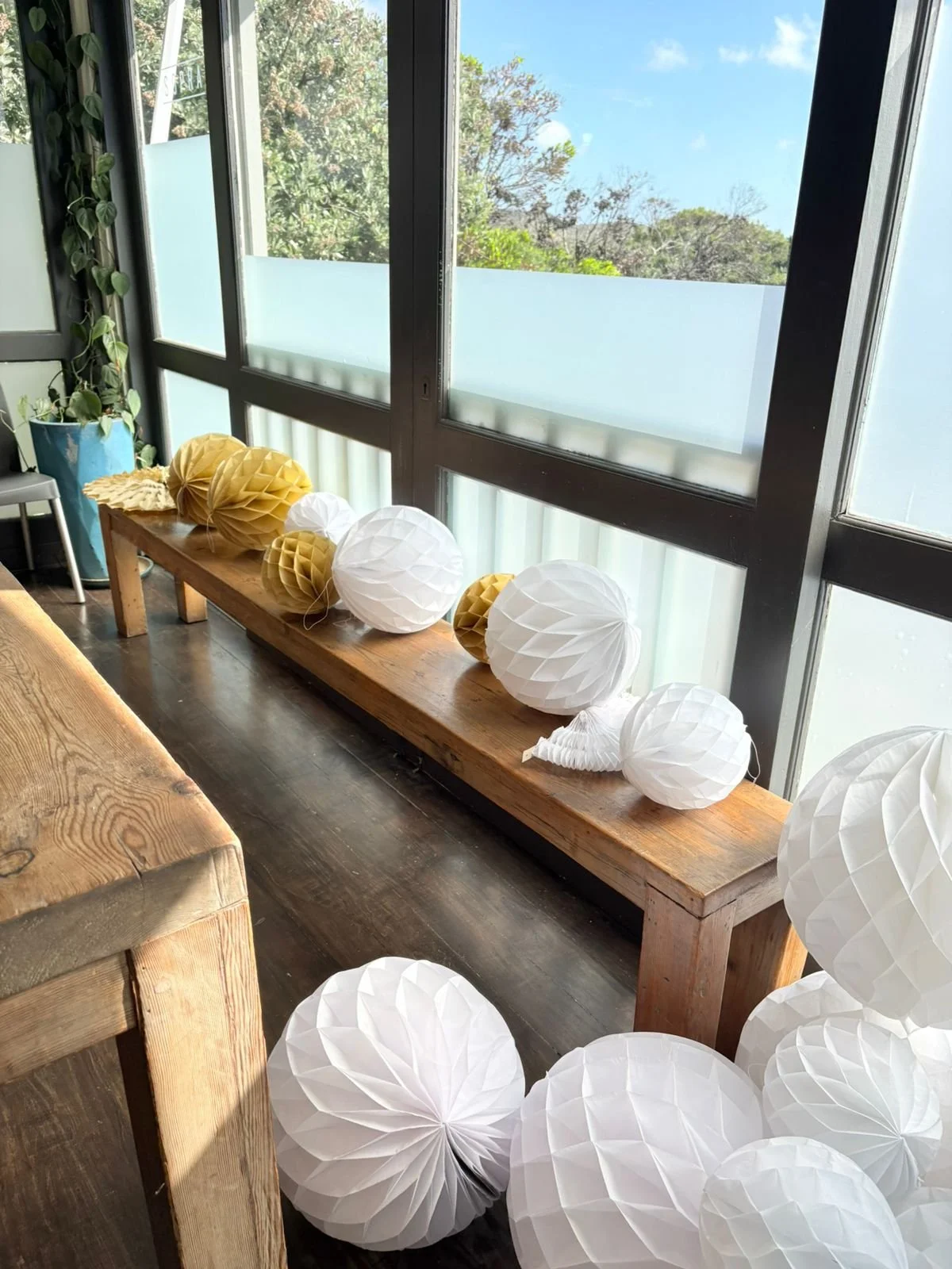 Decorative paper lanterns in white and gold on a wooden bench near large windows with outdoor greenery.