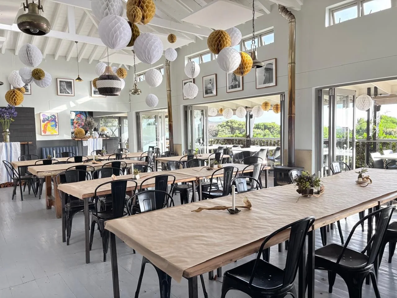 Bright dining area with large windows, decorated with hanging paper lanterns and framed photos on the wall, set for an event with tables covered in beige tablecloths and small floral centerpieces.