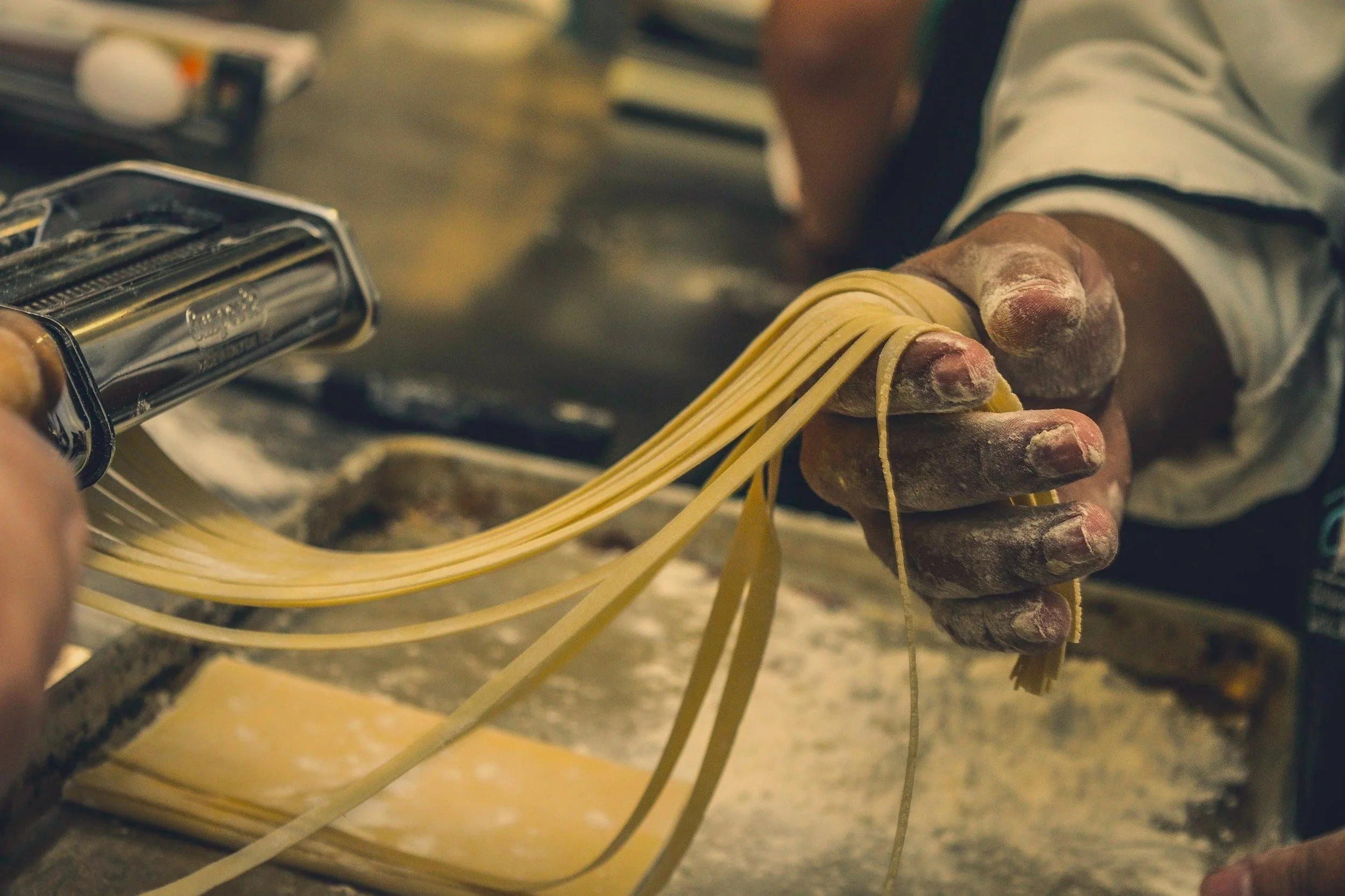 Close-up of a person’s hand pressing dough through a pasta machine, making fresh spaghetti noodles in a kitchen.