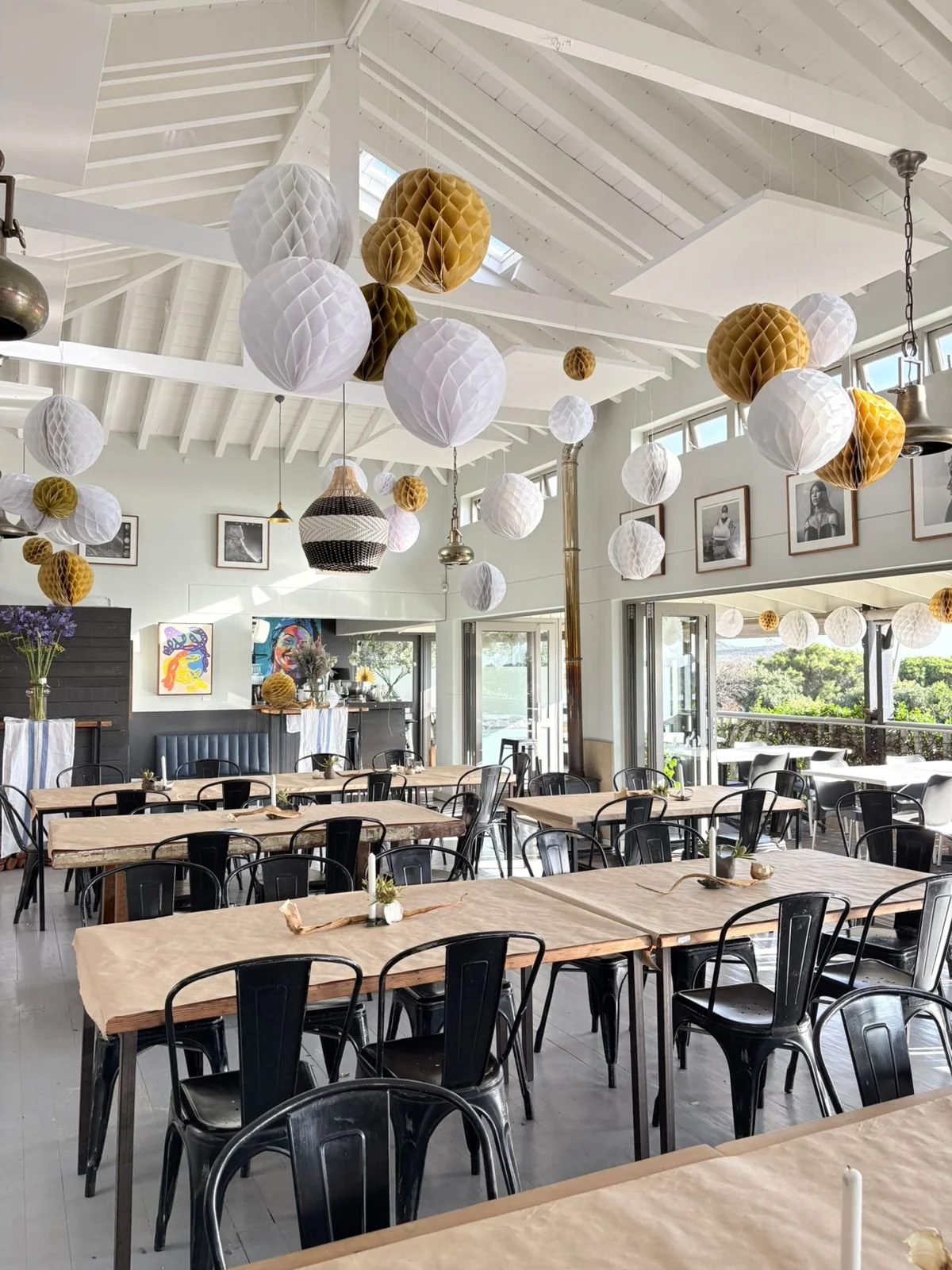 A bright, modern restaurant interior with hanging paper lanterns in white, gold, and brown, wooden tables with beige tablecloths, black chairs, and framed black-and-white photographs on the wall.